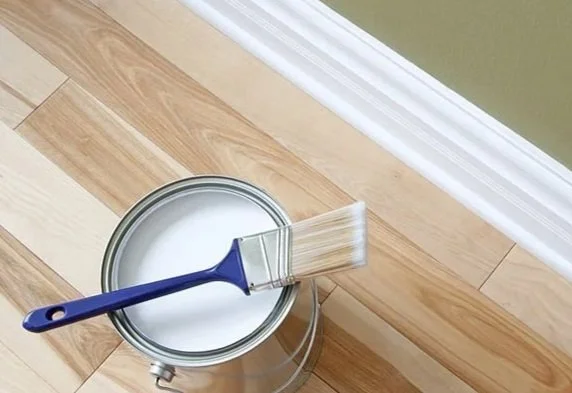 A paint roller with white paint resting inside a small open can of white paint on a wooden floor next to a green wall with white baseboard.