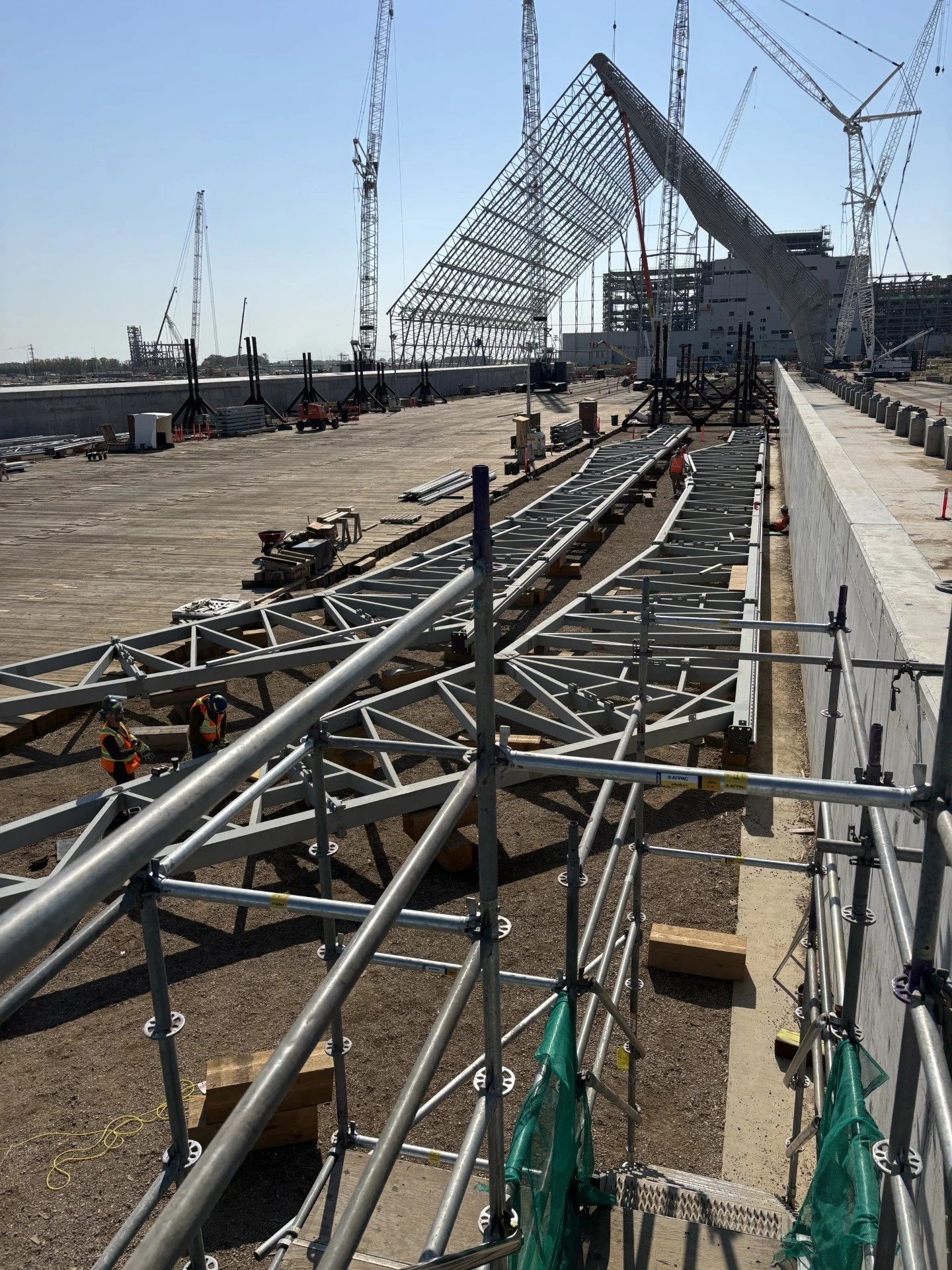 Construction workers on a large construction site assembling a steel framework with cranes in the background, during daytime.