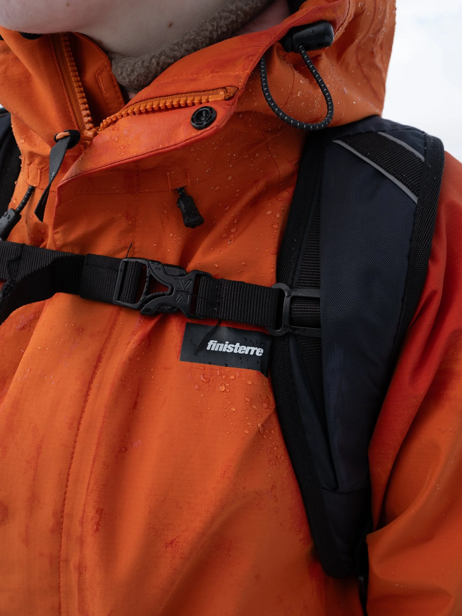 Close-up of a person wearing an orange waterproof jacket and a black backpack with water droplets on the jacket.