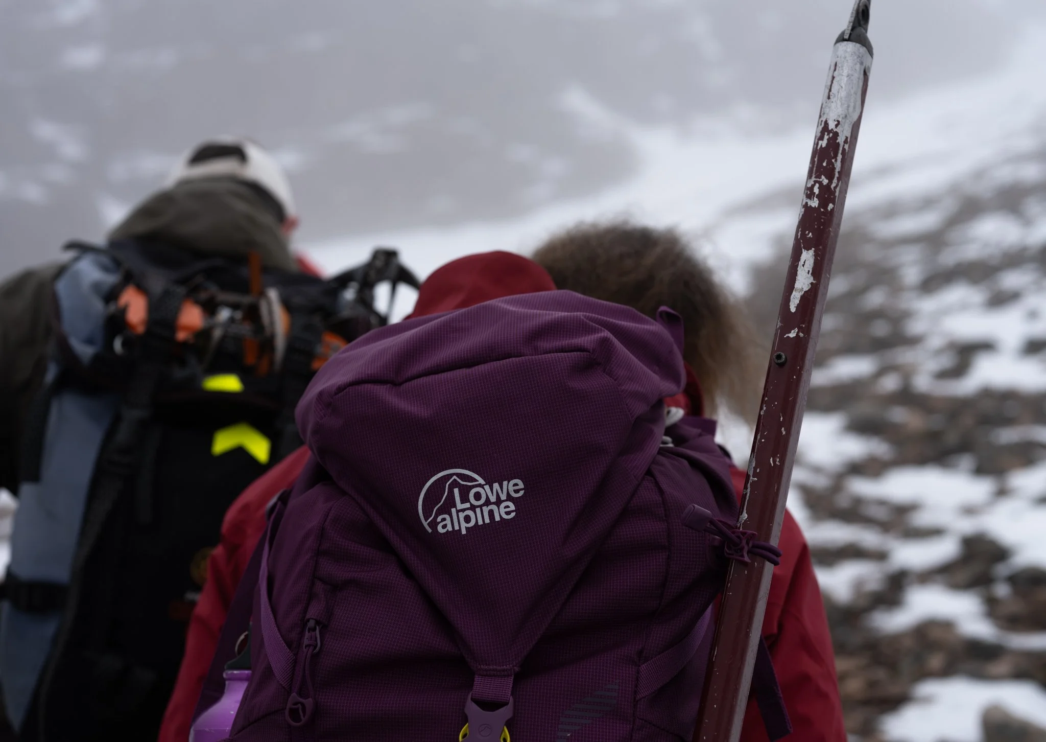 Back view of two hikers climbing snowy mountain terrain, one with a purple Lowe alpine backpack, and the other with a dark backpack, surrounded by snow and cloudy sky.