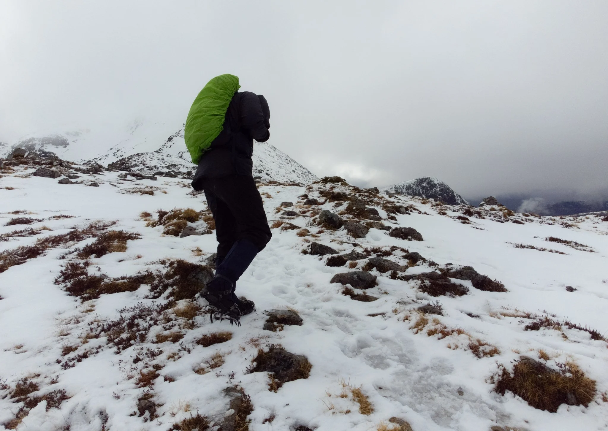 Person hiking on snowy mountain trail with a green backpack, wearing winter gear, cloudy sky in the background.