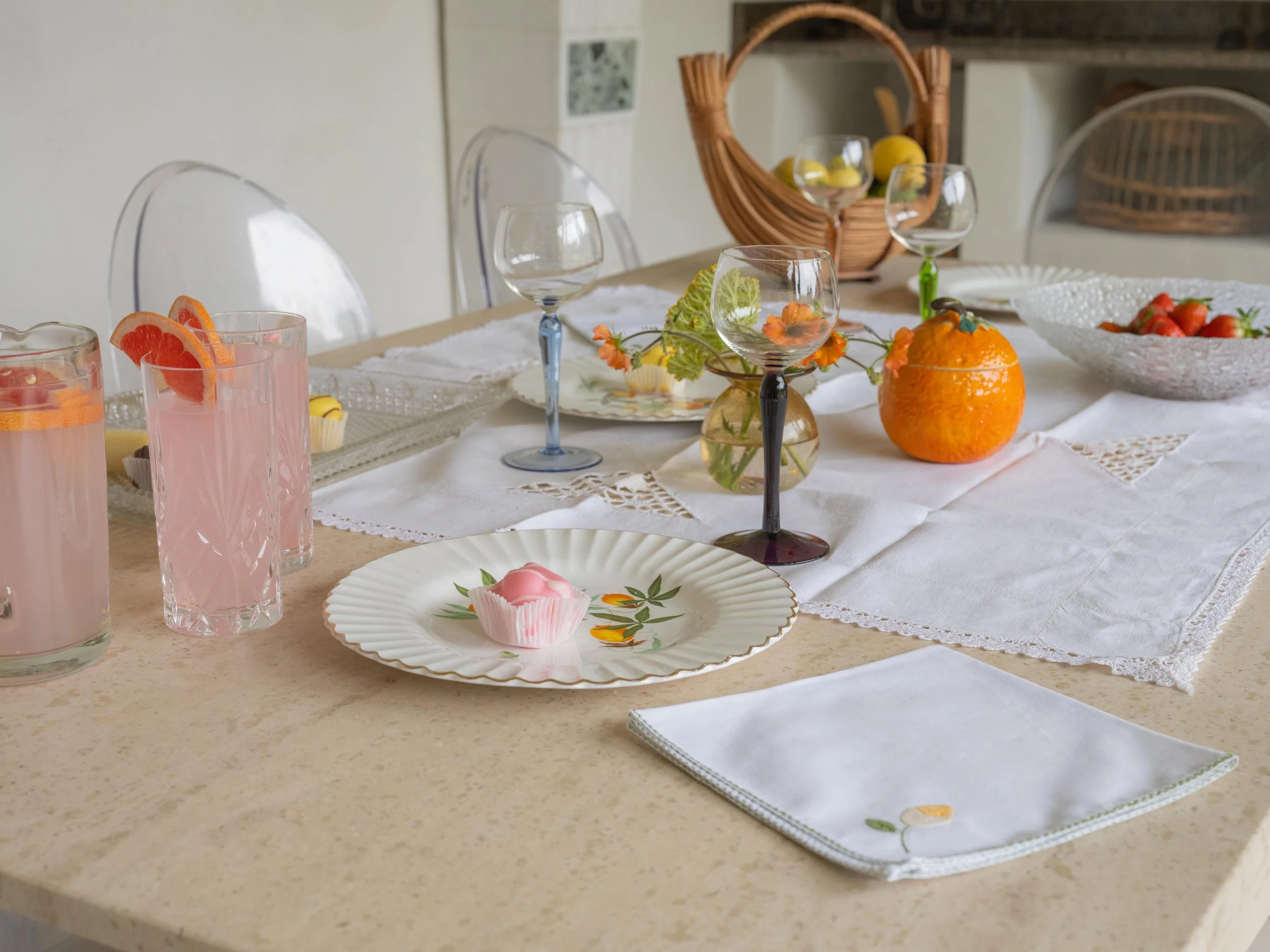 A table set for a meal with white tablecloth, colorful glasses, floral and fruit centerpieces, and dishes including strawberries, oranges, and pink desserts.