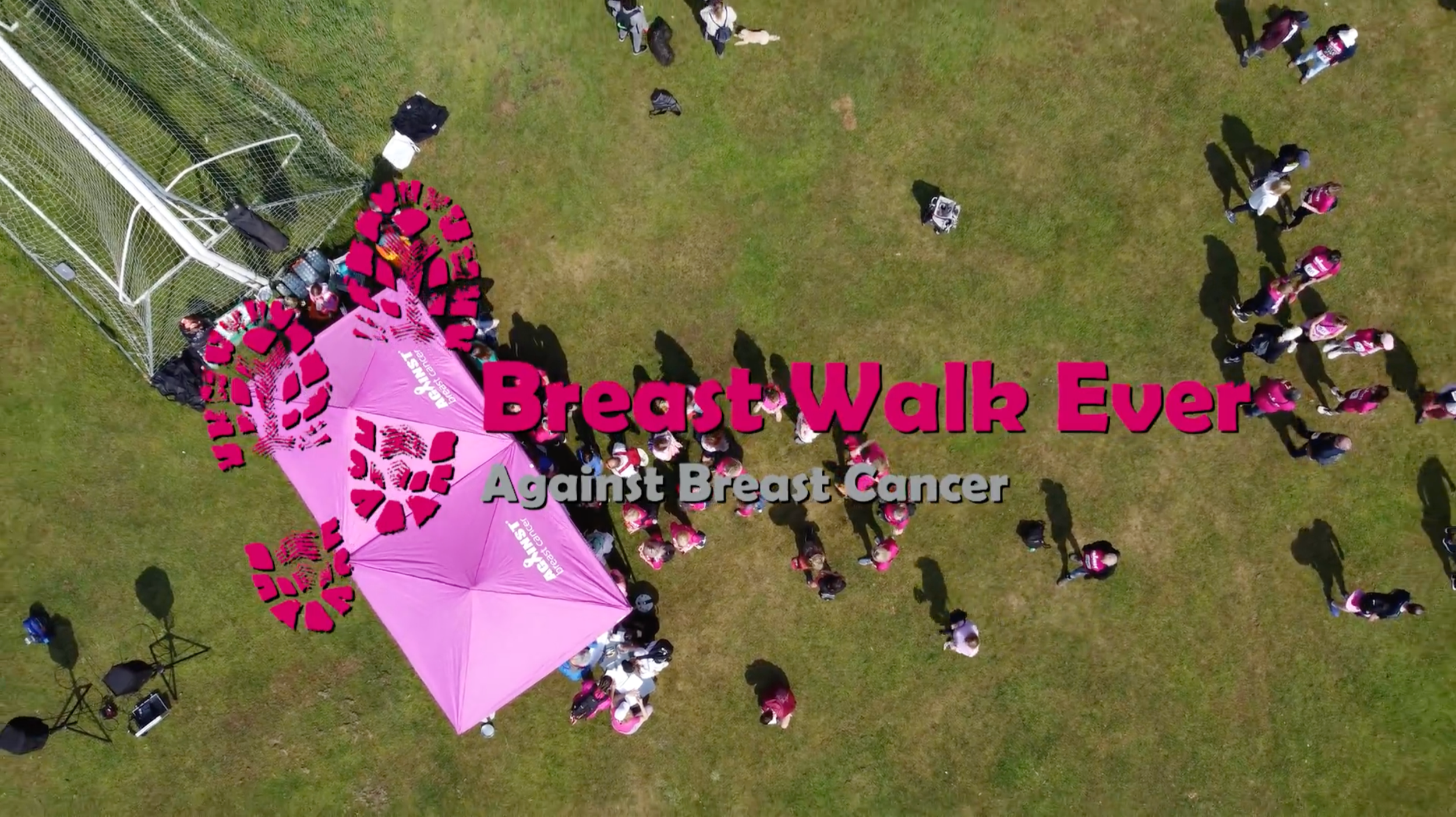 An aerial view of a breast cancer awareness event with a large pink logo and tent, people gathering and walking on a grassy field, supporting breast cancer awareness.