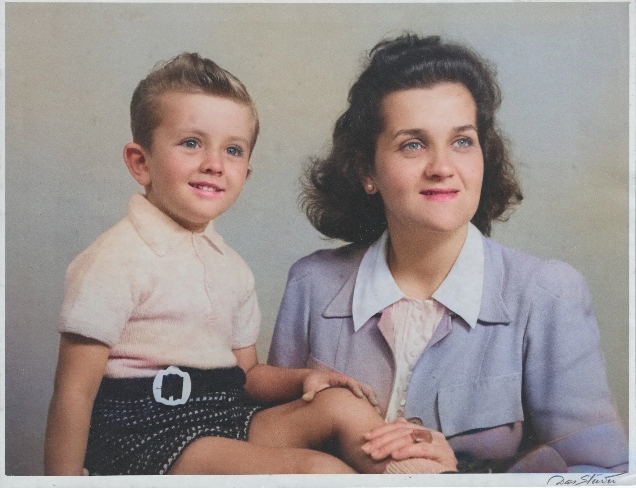 Photograph of a woman with curly dark hair and a young boy with light brown hair, both smiling. The woman is wearing a lavender blazer over a light-colored blouse, and the boy is wearing a light pink shirt and dark shorts with a camera belt.