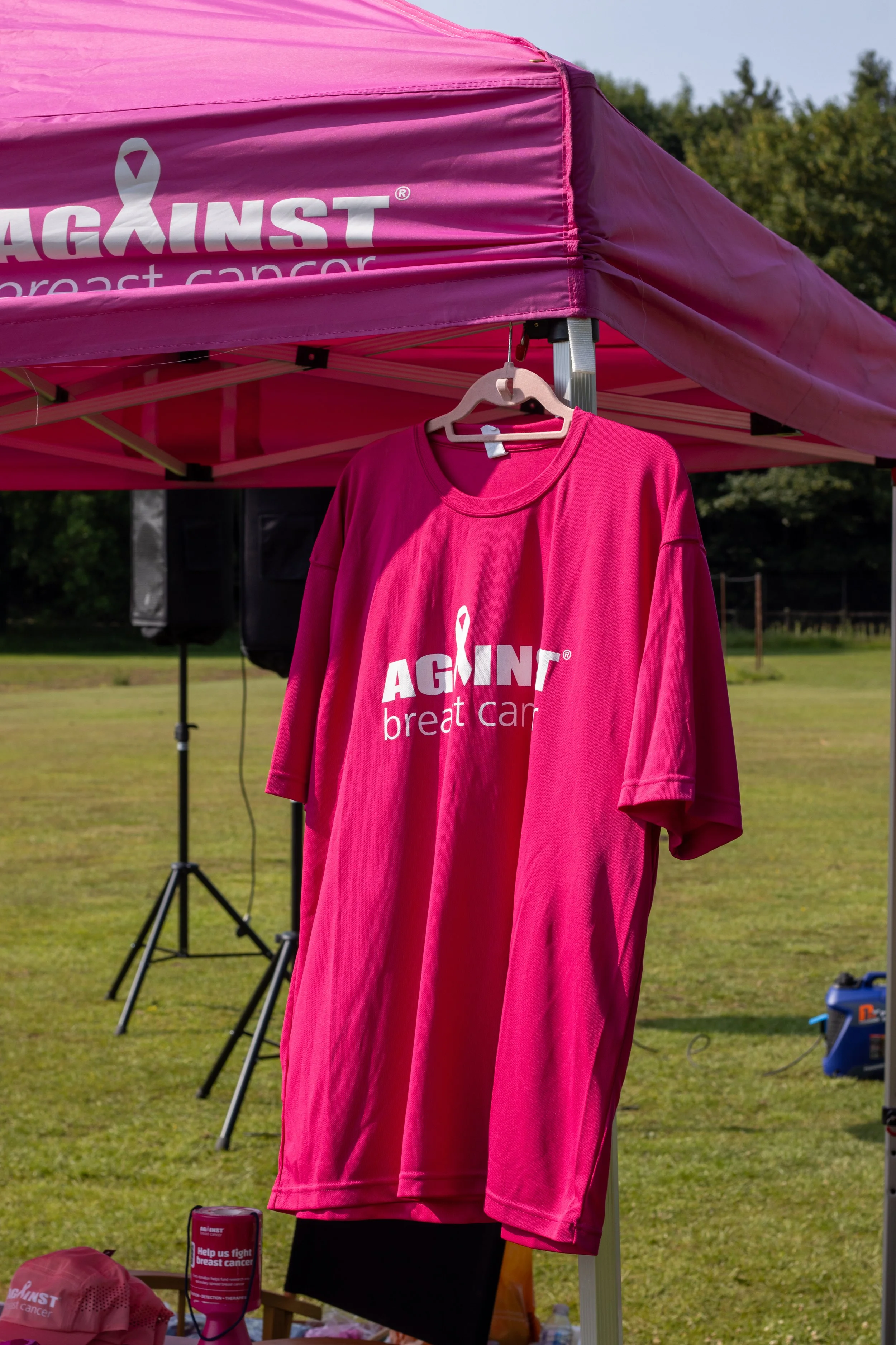 Pink T-shirt hanging under a pink canopy with the logo 'AGAINST breast cancer' on it, set up outdoors on a grassy field.