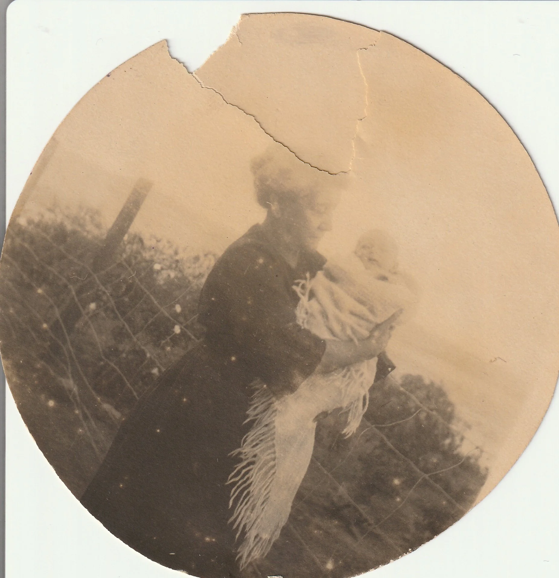 A vintage black and white photograph of a woman holding a dog outdoors, with a wire fence and trees in the background, slightly damaged with a torn top edge.