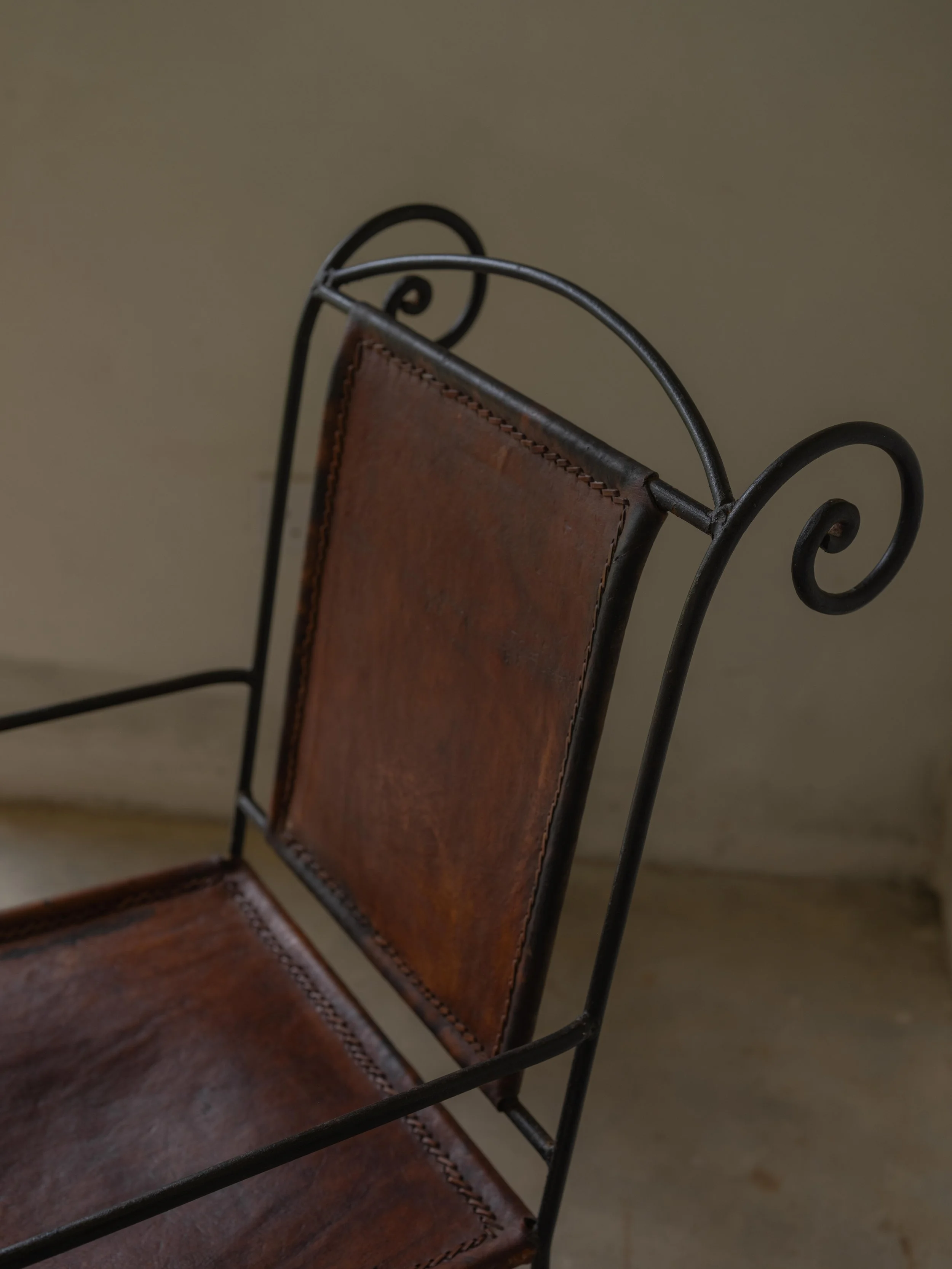 Close-up of a vintage brown leather and black metal chair with scroll details on the armrests, against a plain background.
