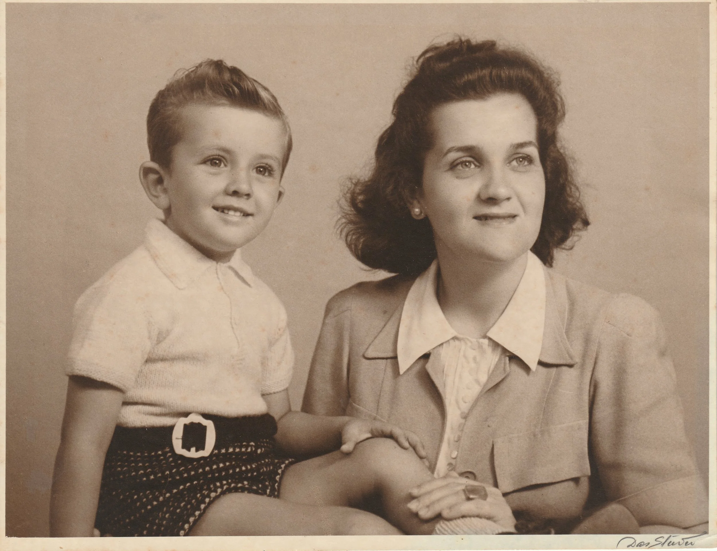 Black and white photo of a woman with curly hair and a young boy with short hair, sitting together and holding hands, both smiling slightly against a plain background.