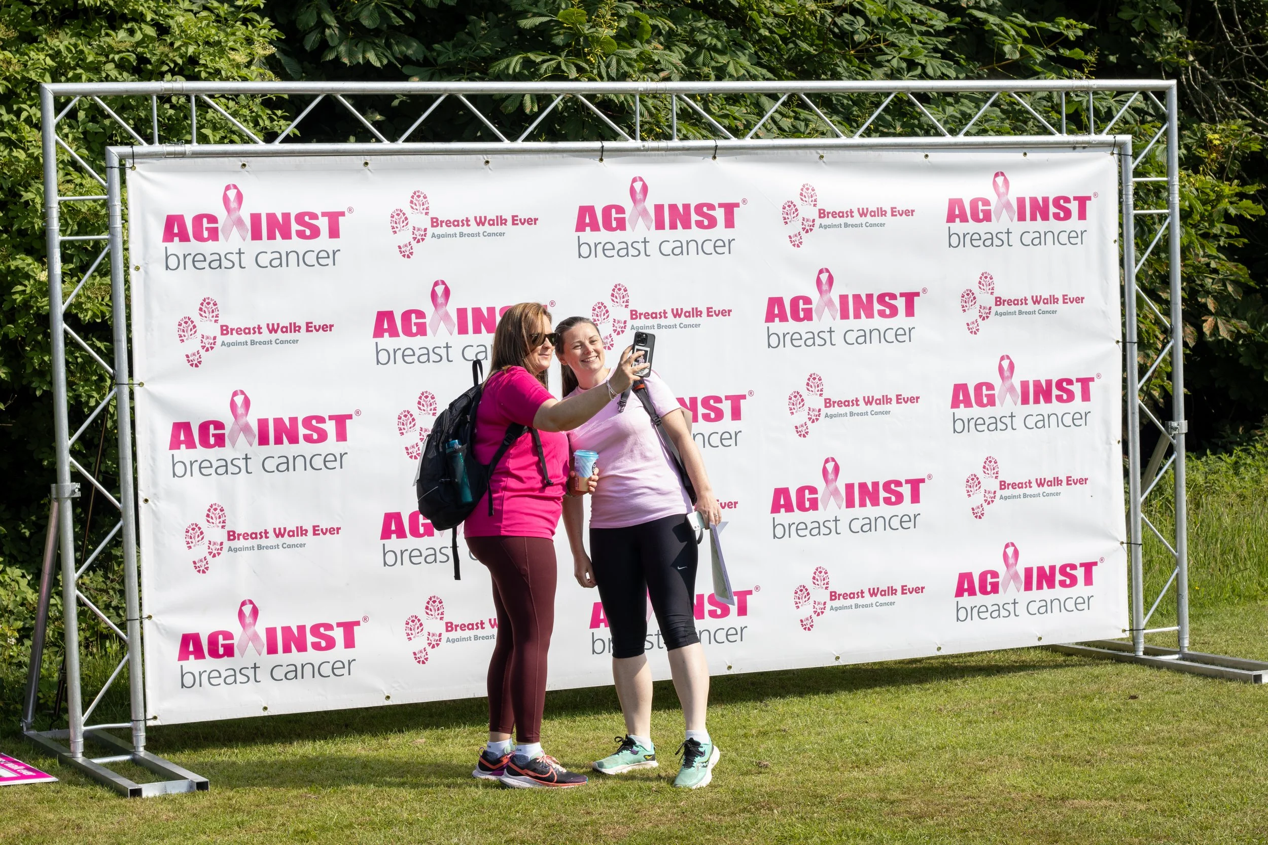 Two women taking a selfie in front of a pink and white Breast Cancer awareness event backdrop. They are dressed in pink and athletic wear, with one wearing sunglasses and a backpack, standing on grass with green foliage behind.