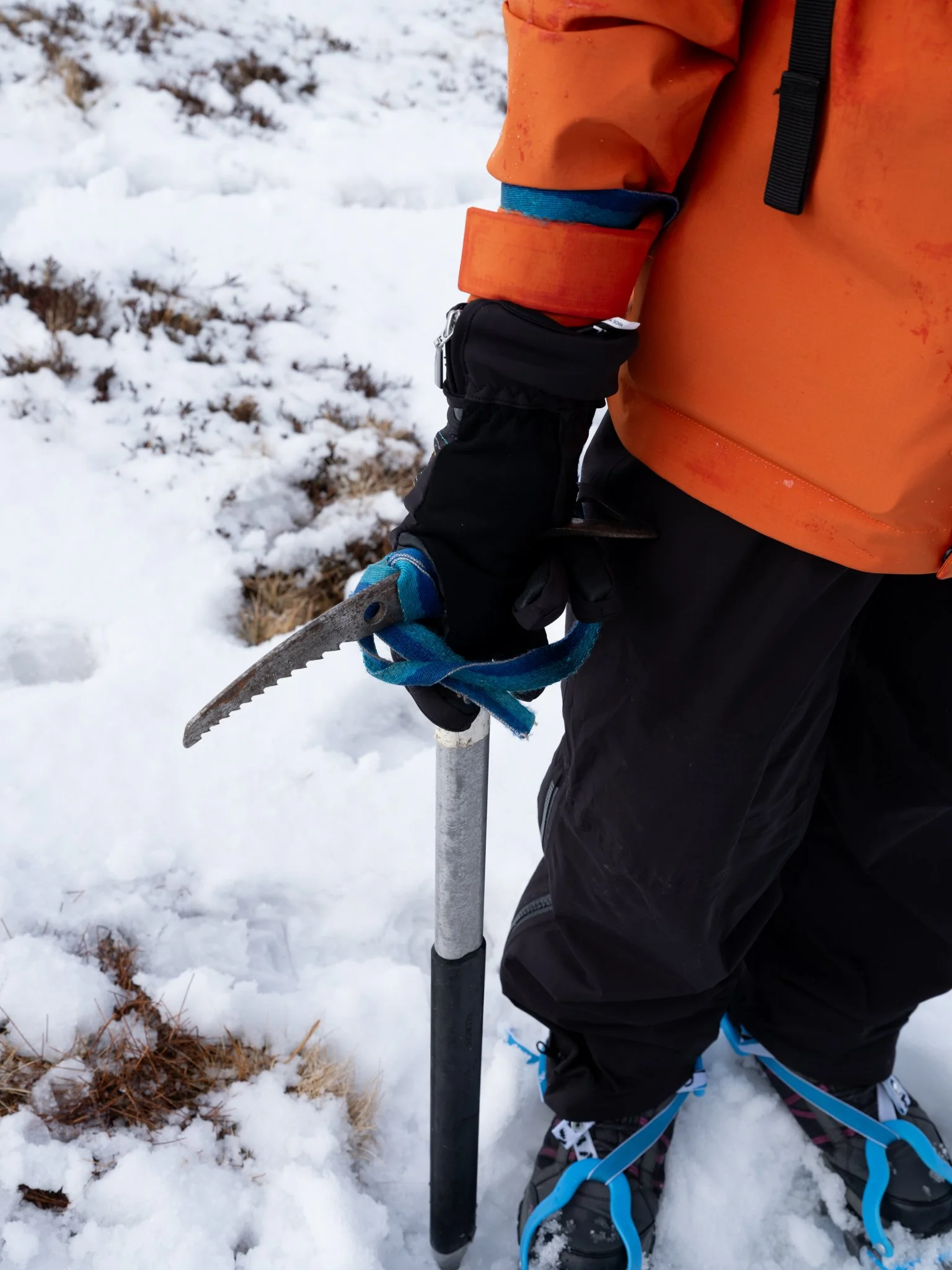 A person dressed in winter outdoor gear, including an orange jacket, black gloves, and snow boots, is standing in snowy terrain holding an ice axe with a blue strap.