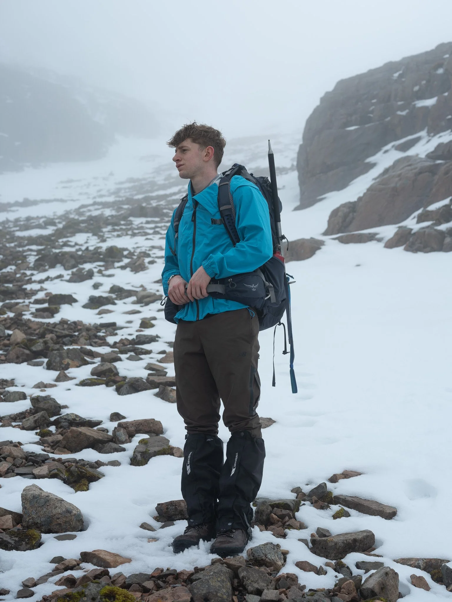 A young man in outdoor gear stands on rocky and snowy terrain in a mountainous area, wearing a bright blue jacket and a large backpack.