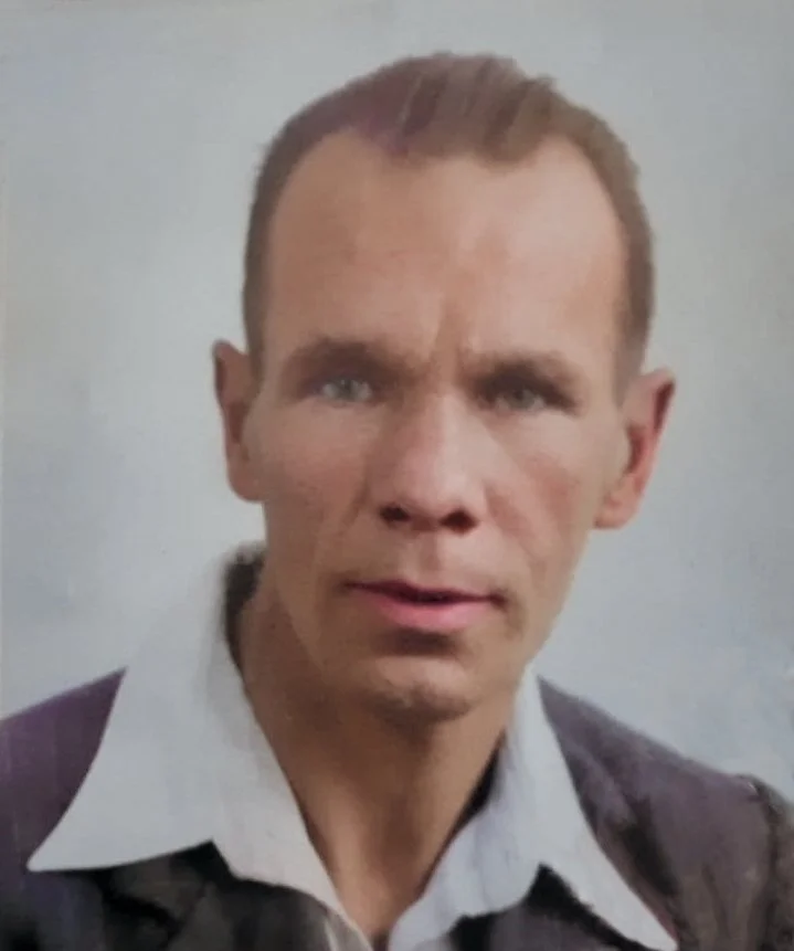 A close-up portrait of a man with short brown hair, wearing a white collared shirt and a dark jacket, against a plain background.