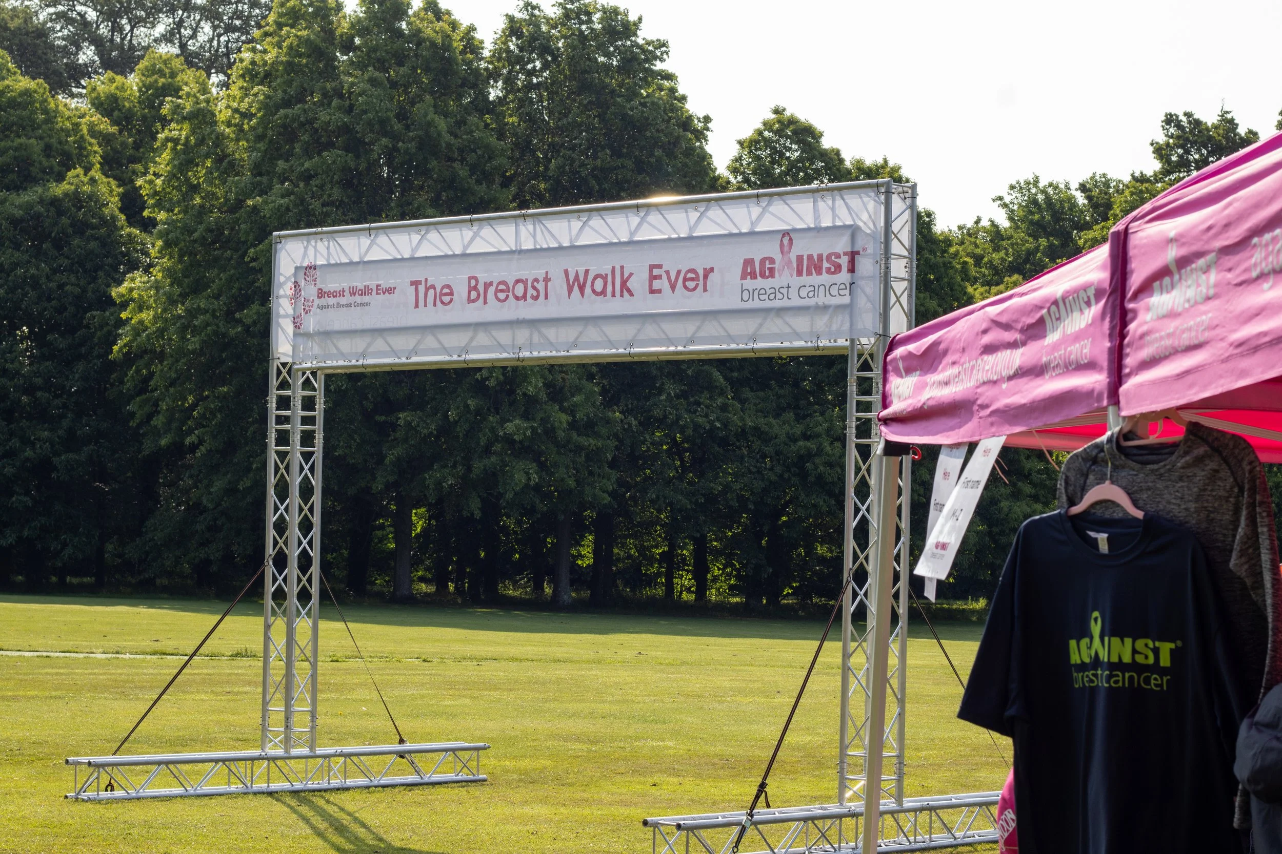 An outdoor setup for a breast cancer awareness walk event with a large banner and nearby pink tents and shirts. The banner reads 'The Breast Walk Ever Against breast cancer' with pink ribbon symbols, set in a grassy area with trees in the background.