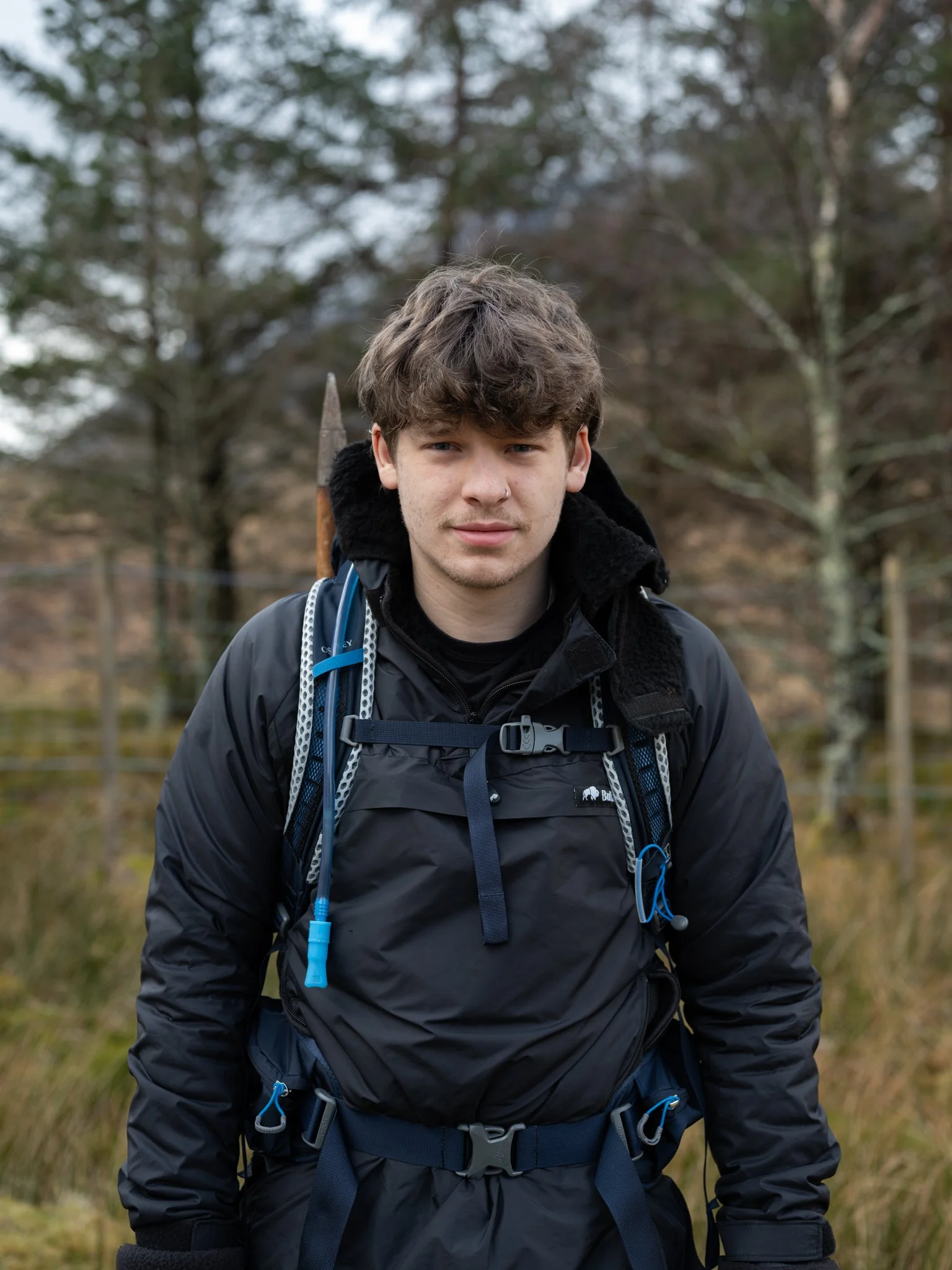 Young man with brown hair, wearing a black hiking jacket and backpack, standing outdoors in a wooded area with trees and grass.