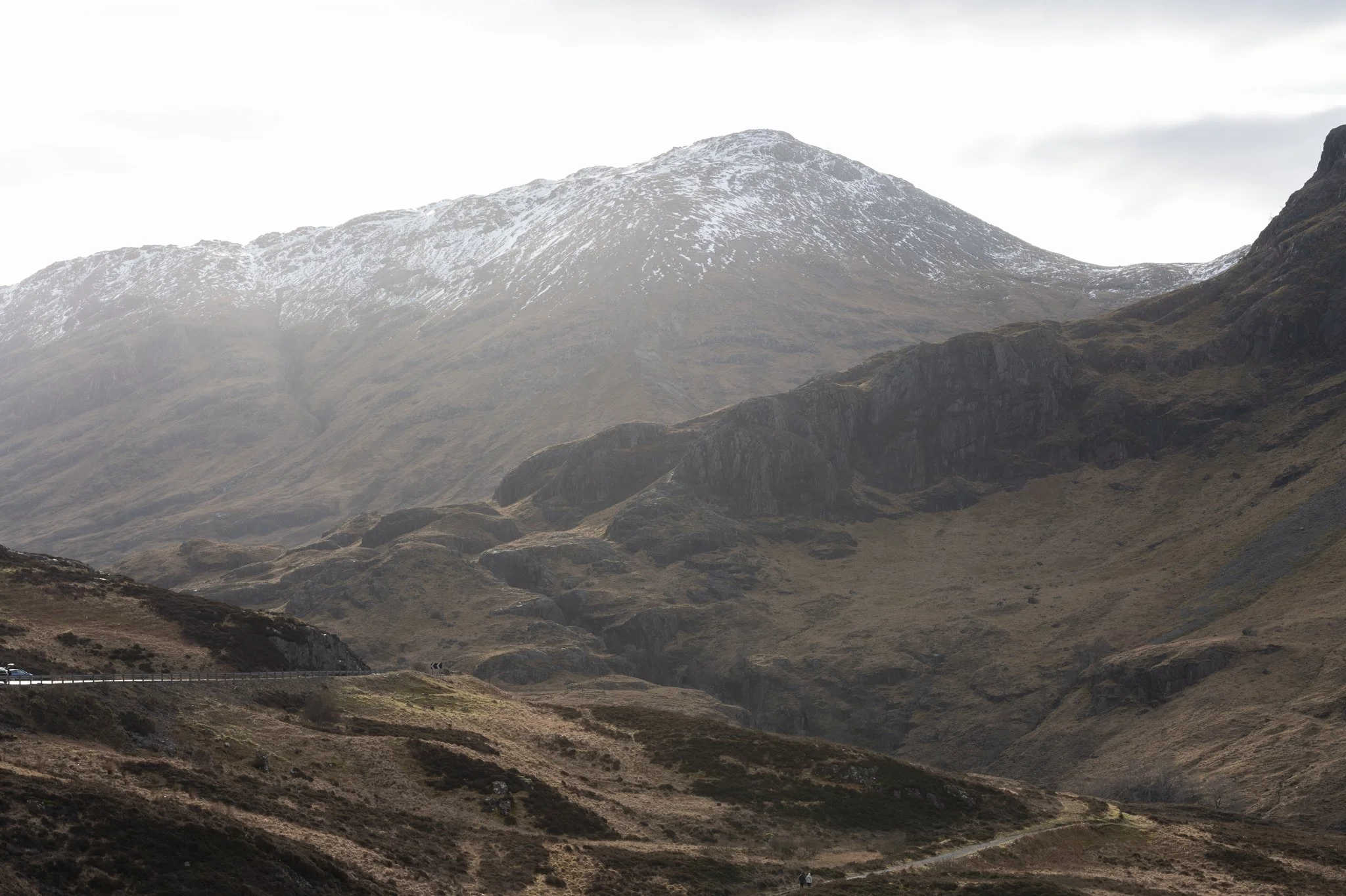 Mountain landscape with snow-capped peak, rugged cliffs, and dry grassy terrain in the foreground.