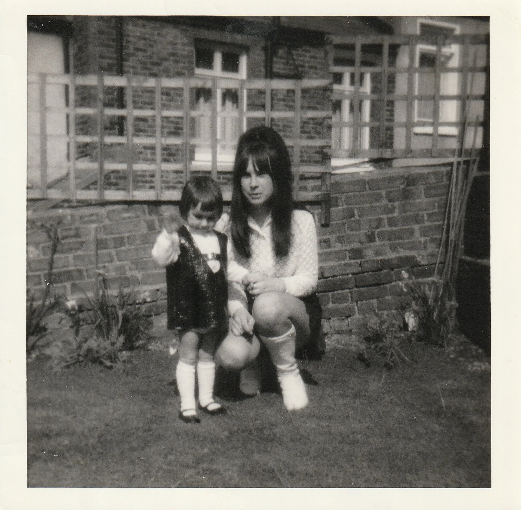 A black and white photo of a woman and a young girl outside in front of a brick wall with a window and a wooden fence. The woman is sitting on the ground, holding the girl's hand, and both are dressed in casual clothing. The girl is wearing a dress, knee-high socks, and sandals, while the woman is wearing a patterned top, skirt, and white socks.