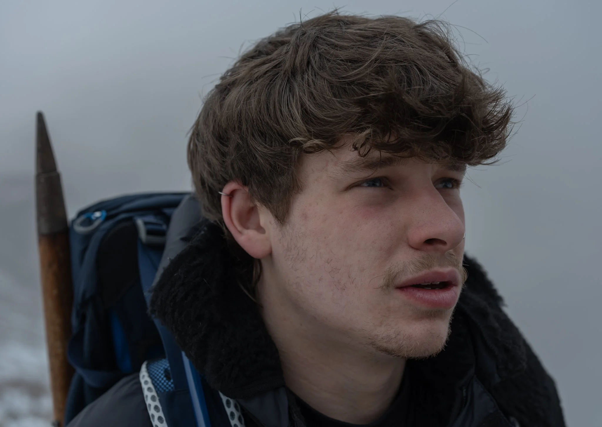 A young man with wavy brown hair and blue eyes looking into the distance, carrying a backpack and a wooden hiking pole, outdoors in a cloudy environment.