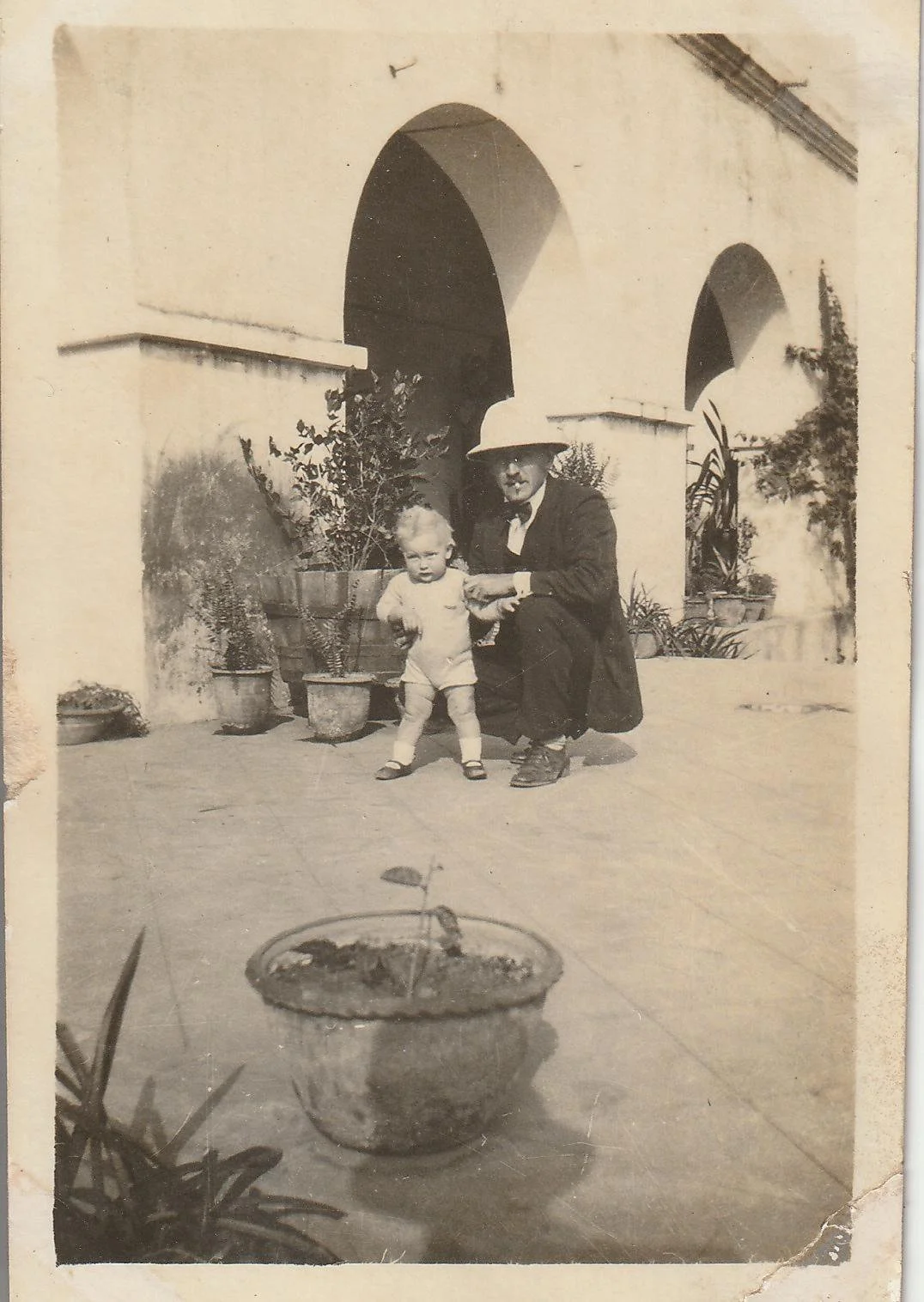 A man with glasses and a wide-brimmed hat crouches next to a small child on a patio with potted plants. The man is dressed in a suit, and the child is in light clothing. The background shows a building with arched doorways.