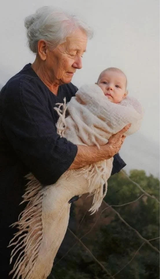 An elderly woman holding a baby wrapped in a cozy blanket outside with a cloudy sky in the background.