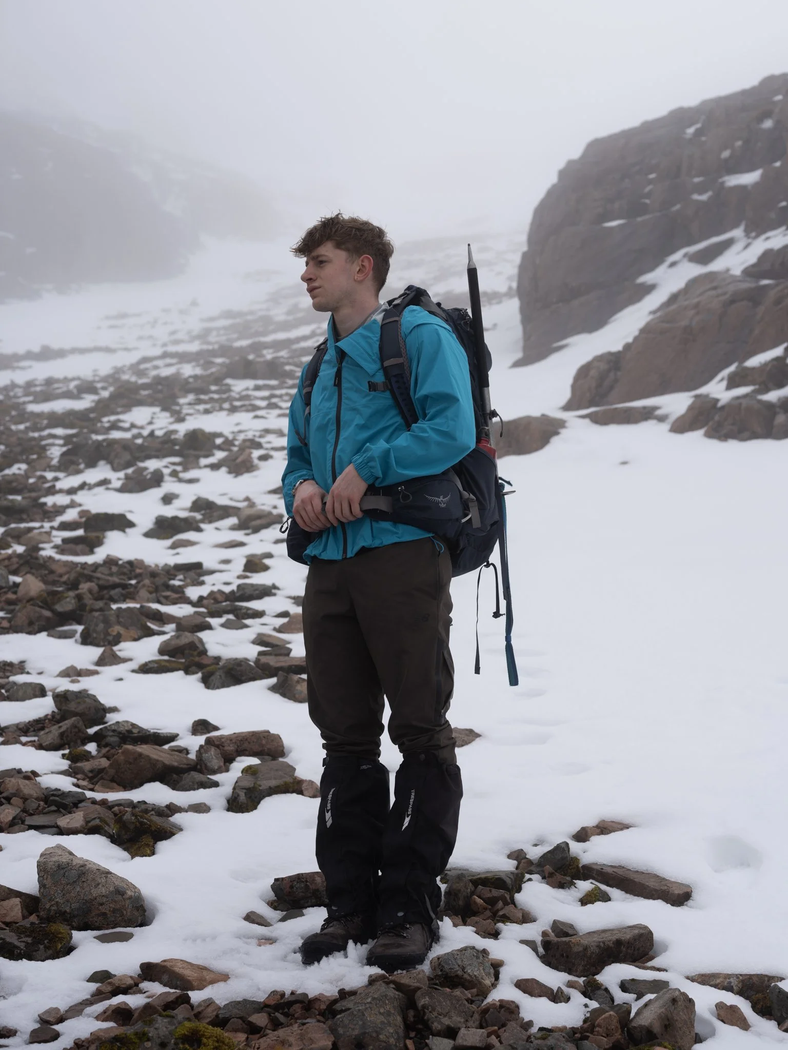 A young man hiking in snowy mountainous terrain, wearing a blue jacket, black hiking pants, and carrying a backpack with hiking gear.
