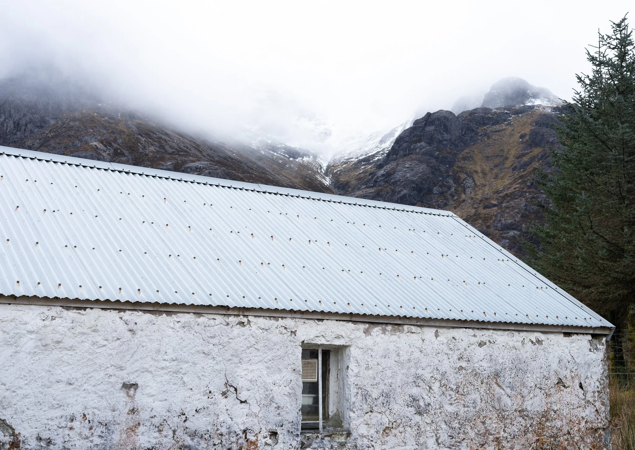 An old white building with a corrugated metal roof in a mountainous landscape, with fog and clouds covering the mountain peaks in the background.