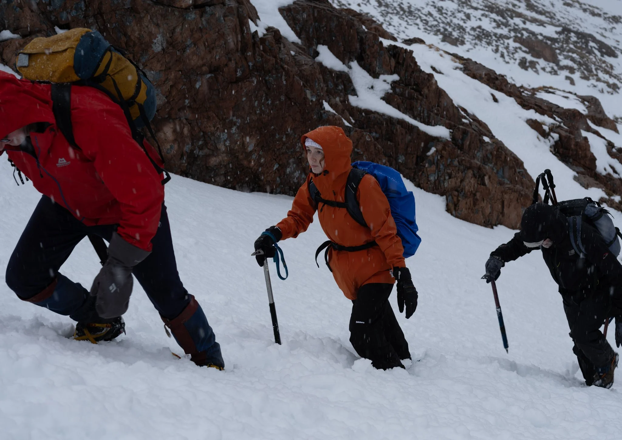 Three climbers in snow, one in a red jacket and two in black, ascending a snowy mountain slope with backpacks, using trekking poles, surrounded by a rocky mountain landscape.