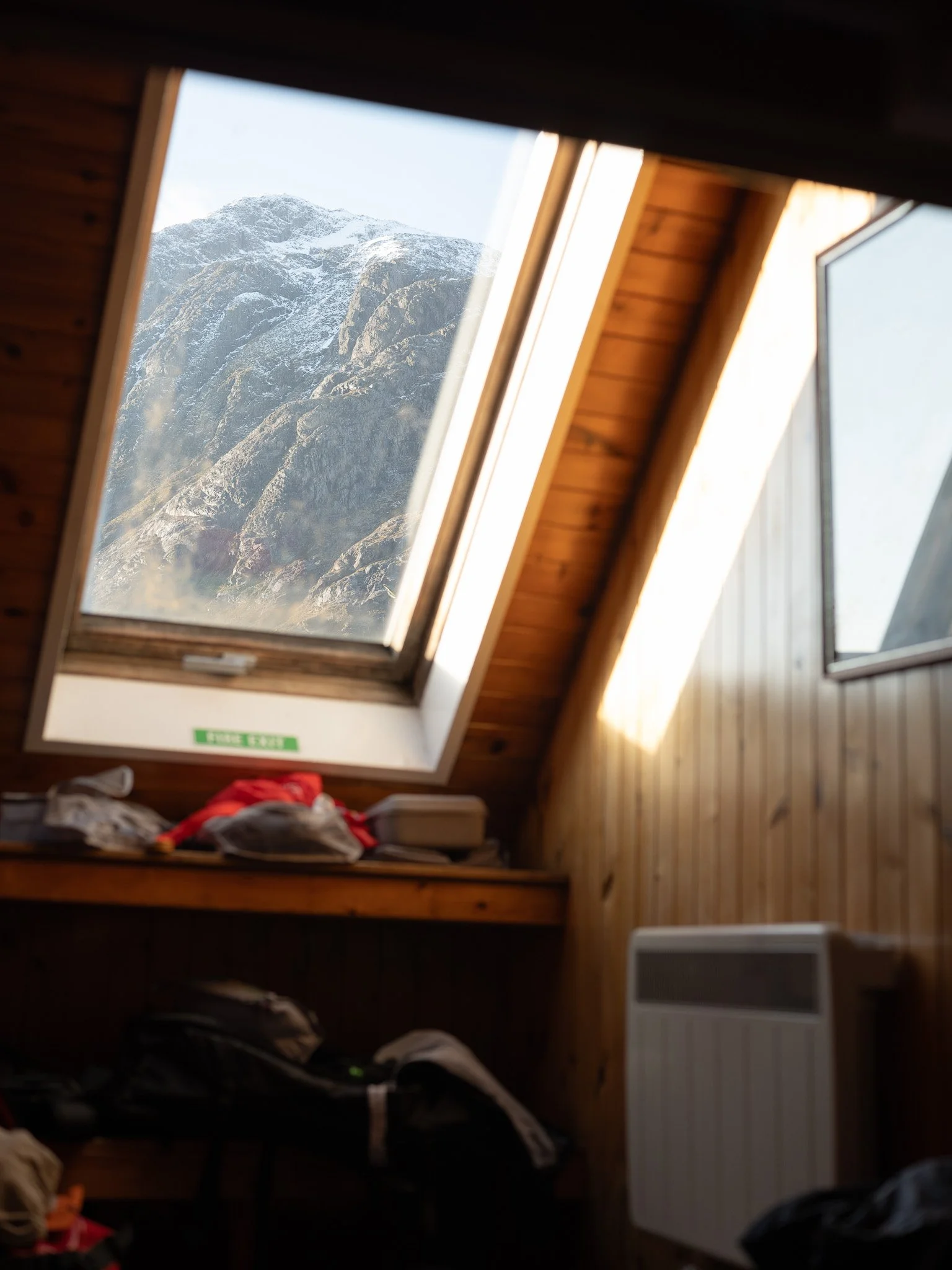 View of a snowy mountain through a skylight window in a wooden attic room, with some clothing and objects on the window ledge and a heater below.