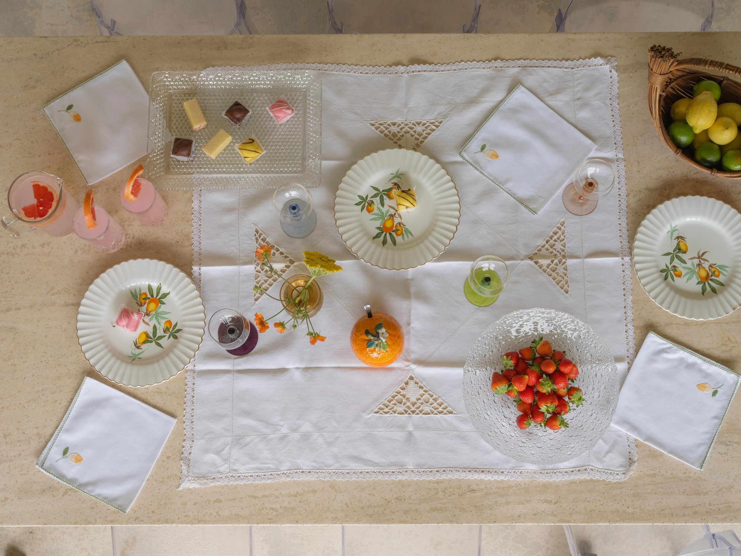 A top-down view of a table setting with white embroidered napkins, six glass cups filled with colorful drinks and garnished with fruit, three decorative plates with peaches and oranges, a glass plate with strawberries, a basket of limes and lemons, and a small vase with yellow and orange flowers, all placed on a white tablecloth with embroidered triangles.