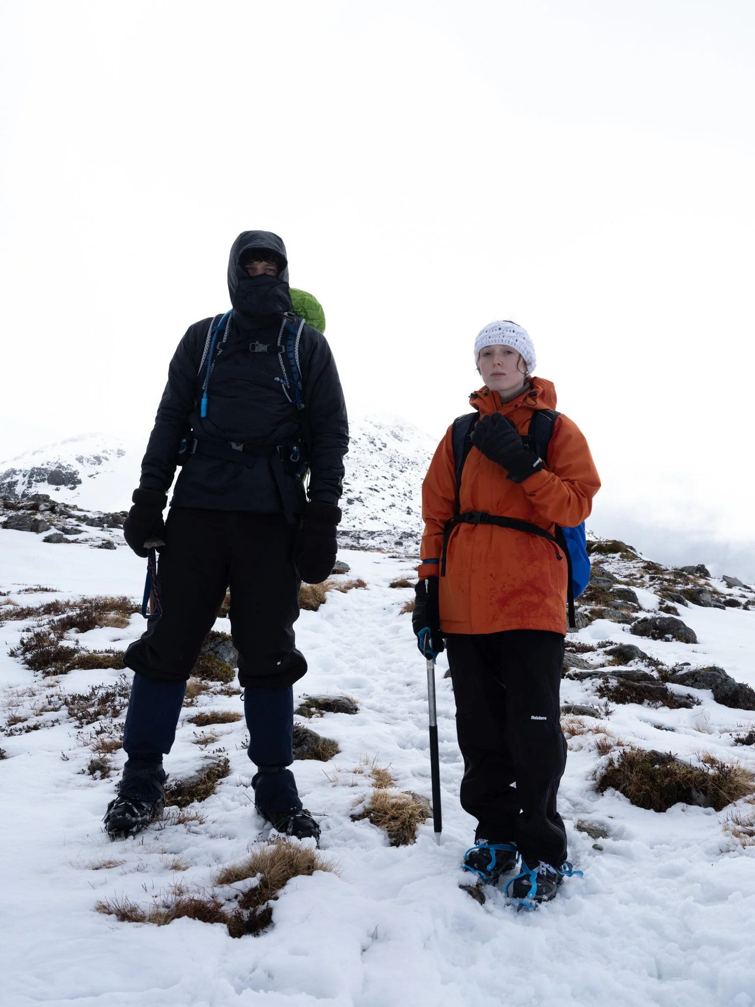 Two people standing in snowy mountainous terrain dressed in winter hiking gear, with one holding a trekking pole.