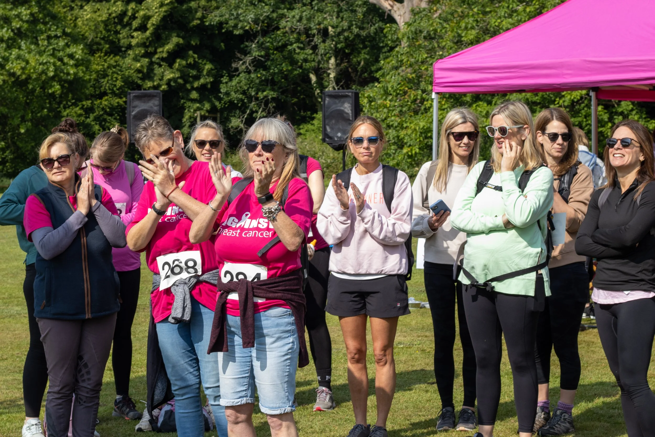 Group of women gathered outdoors at a breast cancer awareness event, some wearing pink shirts with breast cancer awareness logos, standing near a pink canopy, with trees in the background.