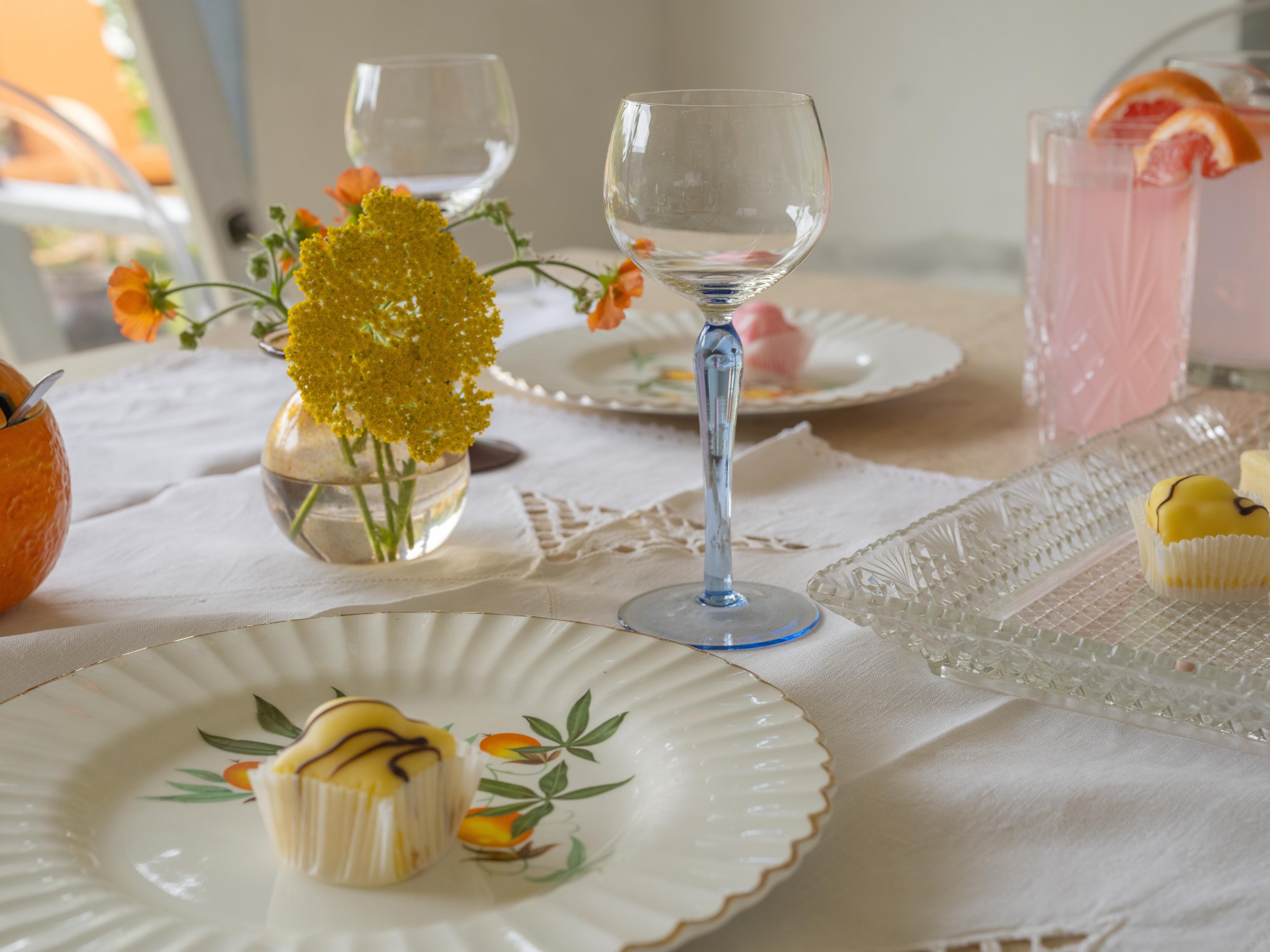 Elegant table setting with a clear glass of wine, a small vase with yellow flowers, a plate with a decorated cupcake, and pink strawberry drinks with orange slices on a glass pitcher, on a white tablecloth.