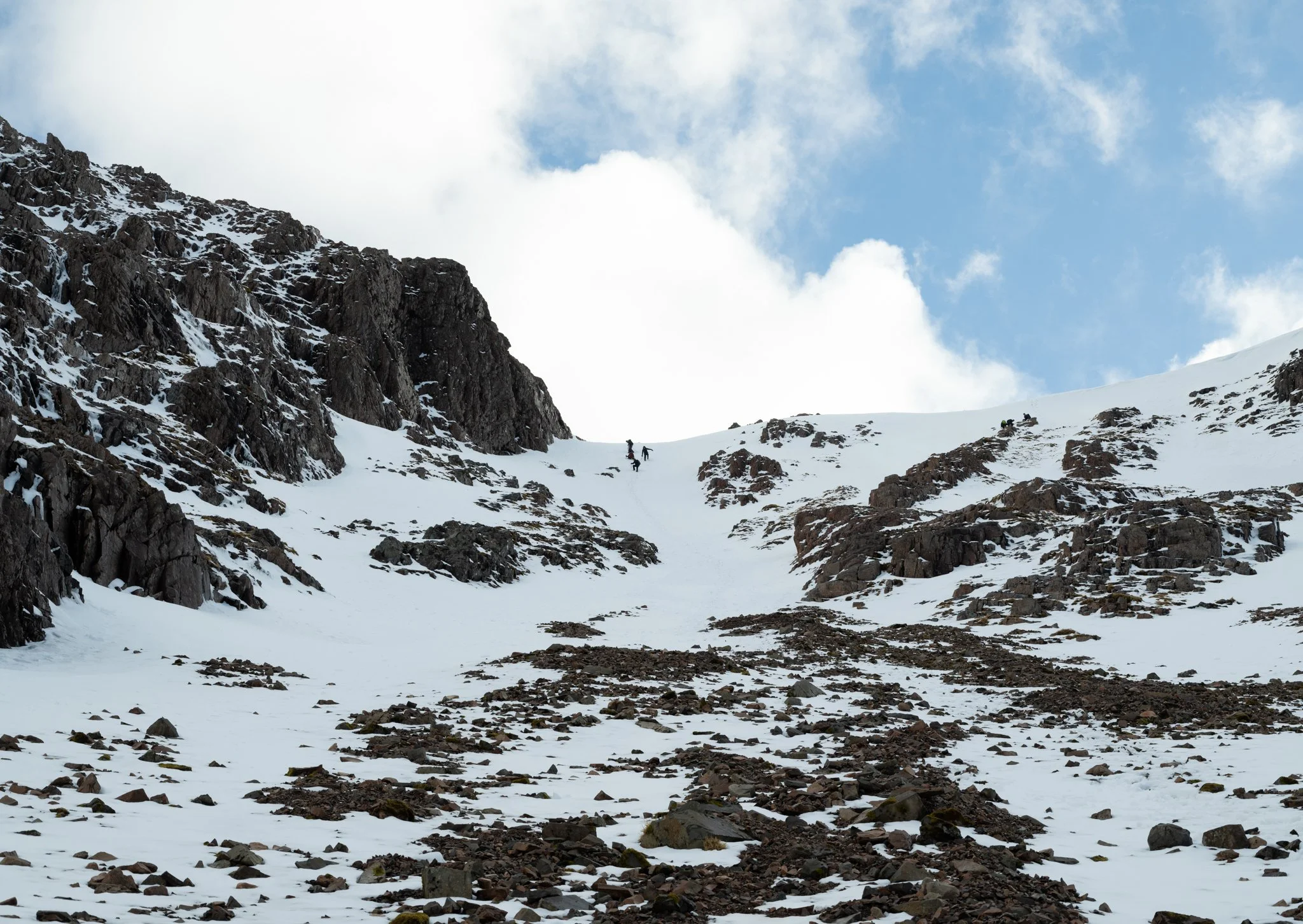 Snow-covered mountain slope with rocky terrain and two hikers ascending, against a partly cloudy sky.