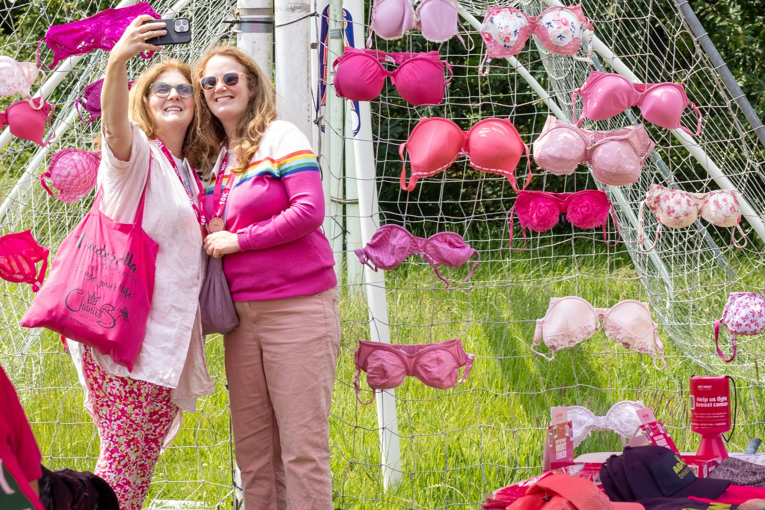 Two women taking a selfie in front of a display of pink and white bras at a breast cancer awareness event, with pink ribbons and donation items nearby.
