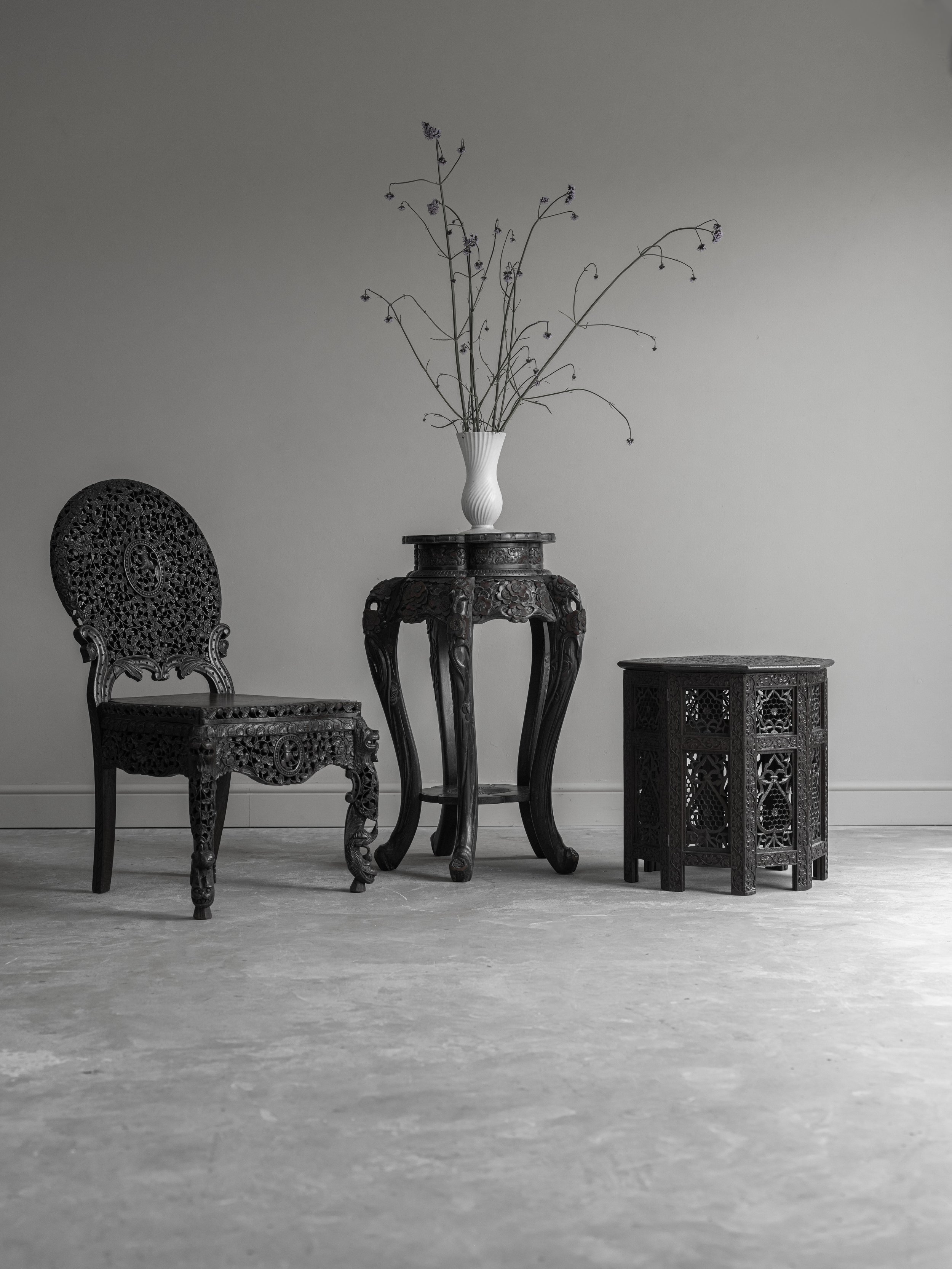 Black carved wooden furniture including a chair, table, and side cabinet, with a white vase and dried flowers on the table, against a plain gray wall.