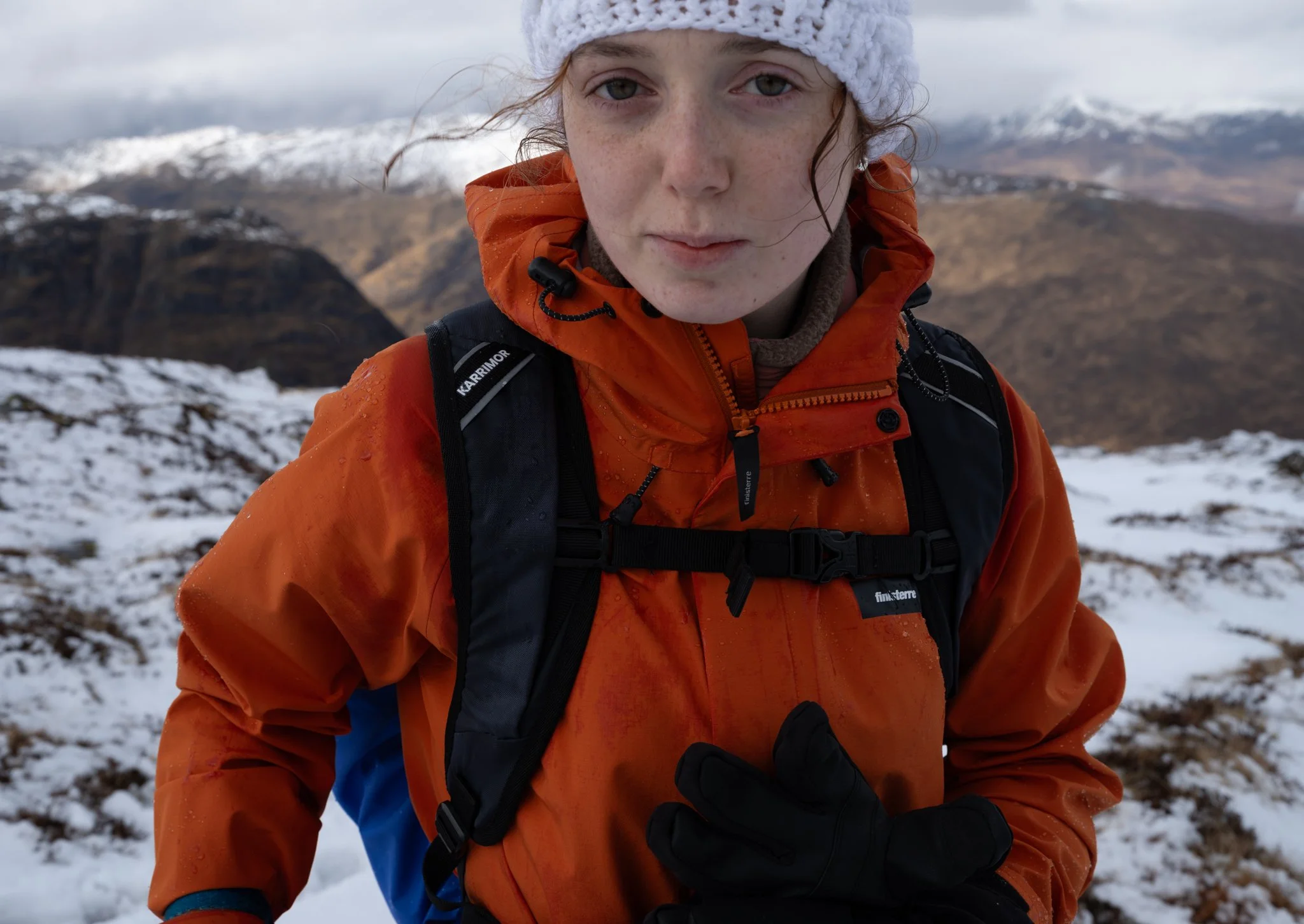 A woman dressed in an orange waterproof jacket and black gloves, carrying a backpack, hiking in a snowy mountainous landscape with cloudy skies.
