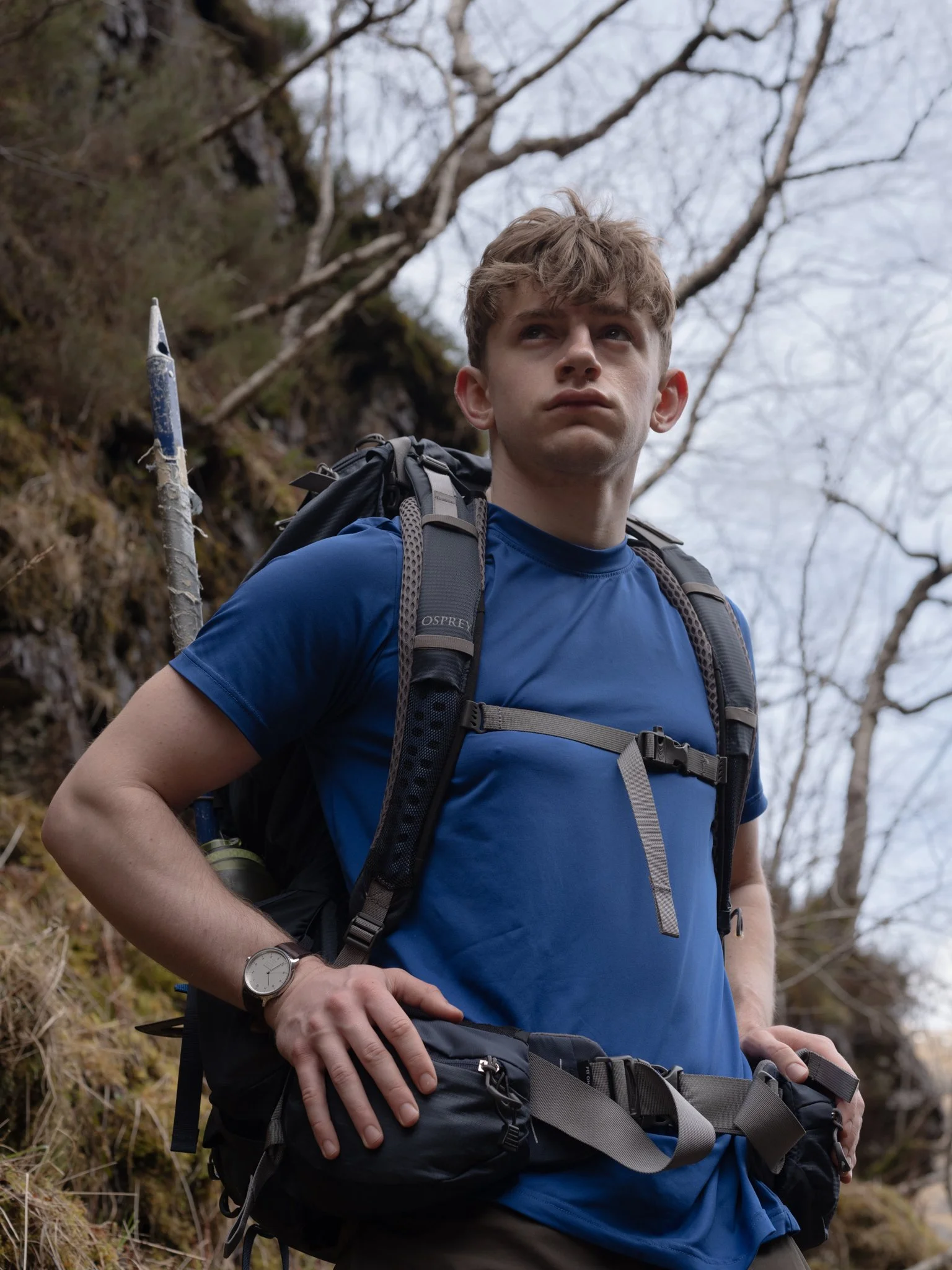 A young man with a backpack hiking outdoors on a trail surrounded by trees and rocky terrain.