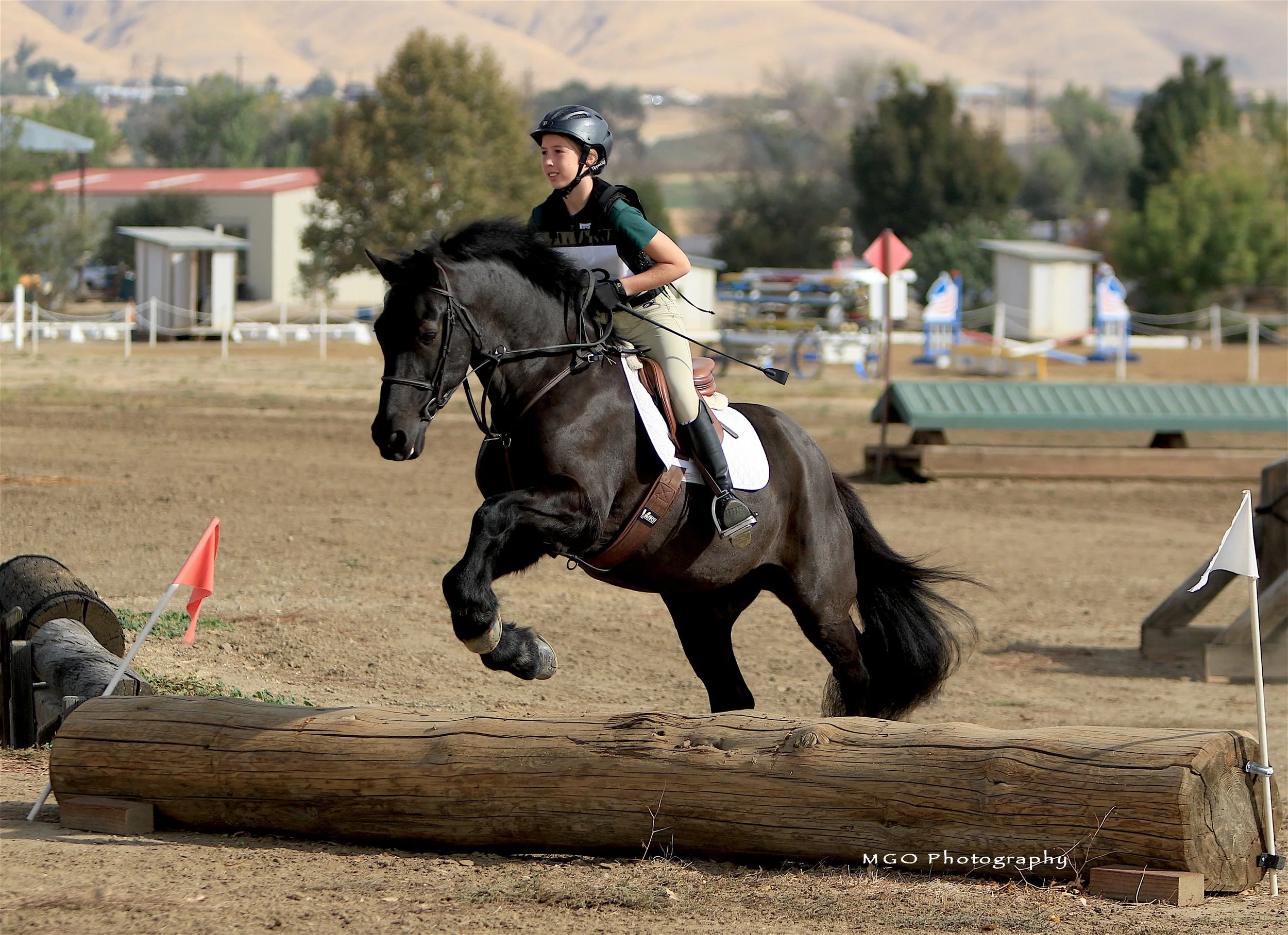 A young girl riding a black horse jumps over an obstacle on a dirt arena during a horse jumping competition. The girl is wearing a helmet, beige pants, a dark green shirt, and riding boots.