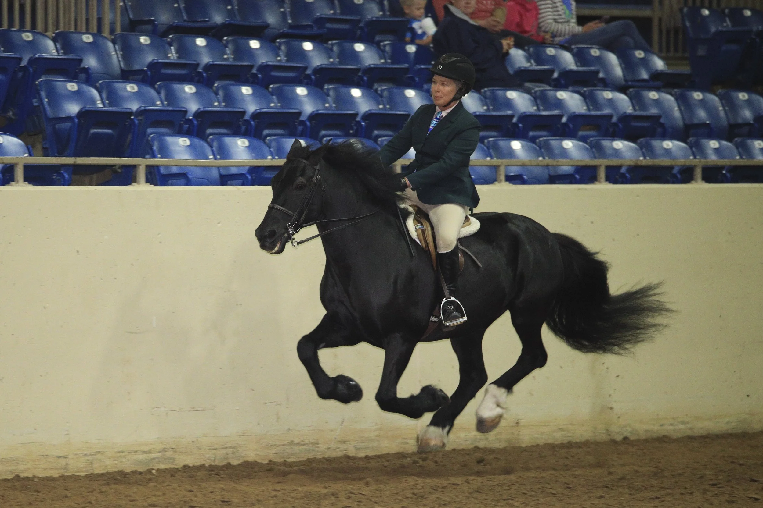 A woman wearing a black helmet, green jacket, and beige riding pants riding a black horse with flowing tail in an indoor equestrian arena. There are blue stadium seats and a few spectators in the background.