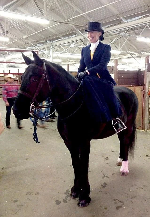 A woman riding a black horse sitting side saddle, in an indoor barn, dressed in formal equestrian attire with a top hat and jacket, smiling.
