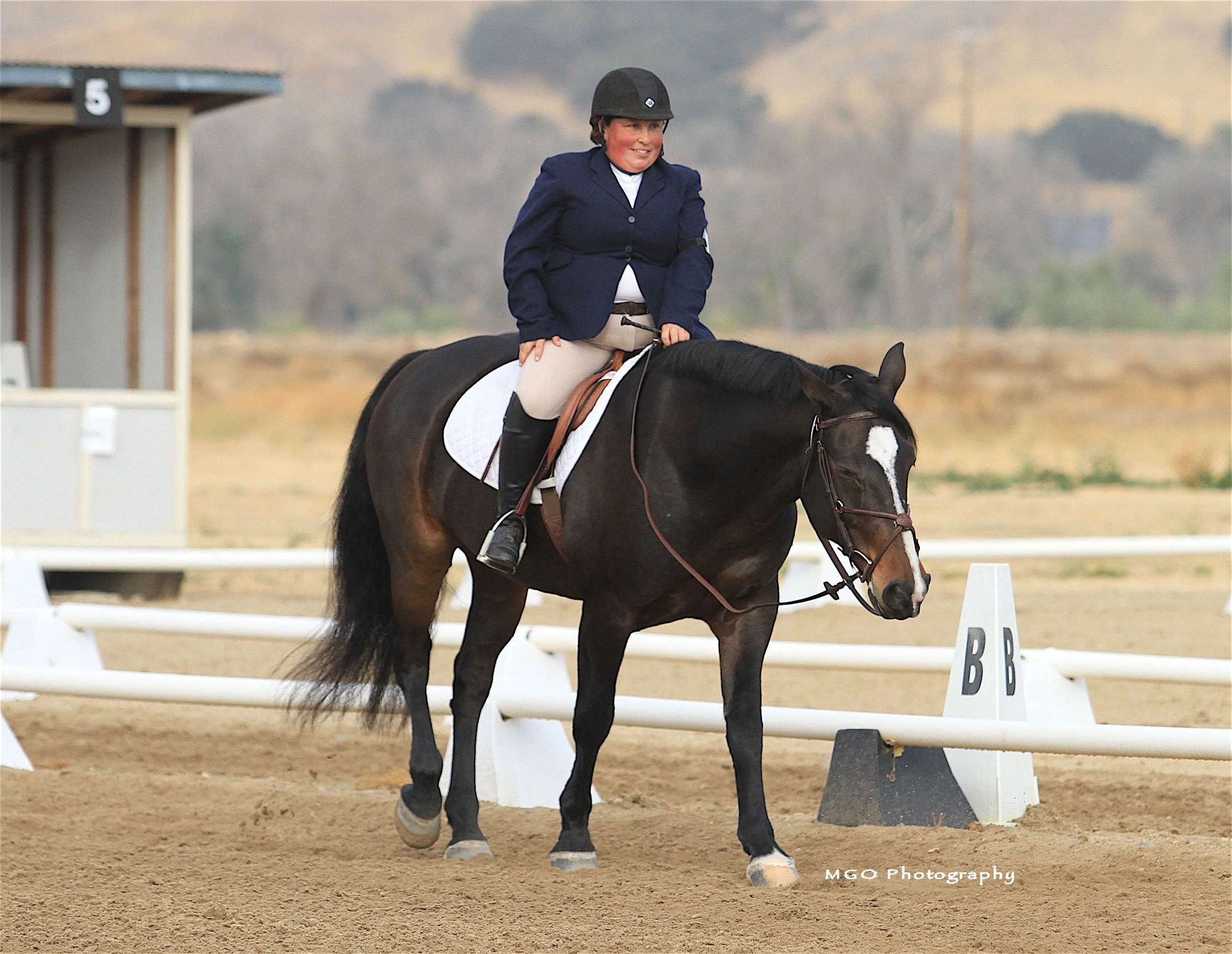 A woman riding a black horse on a dirt horse-riding arena with white railings and dressage markers, wearing a navy jacket, beige riding pants, and a helmet.