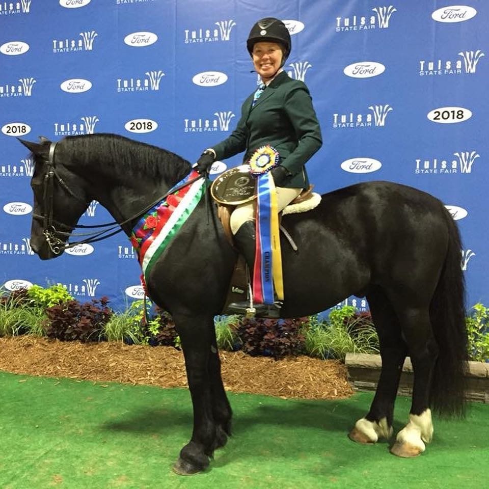 A woman in equestrian attire sitting on a black horse, which has ribbons and a trophy, in front of a backdrop with Tulsa State Fair and Ford logos, celebrating a victory in 2018.