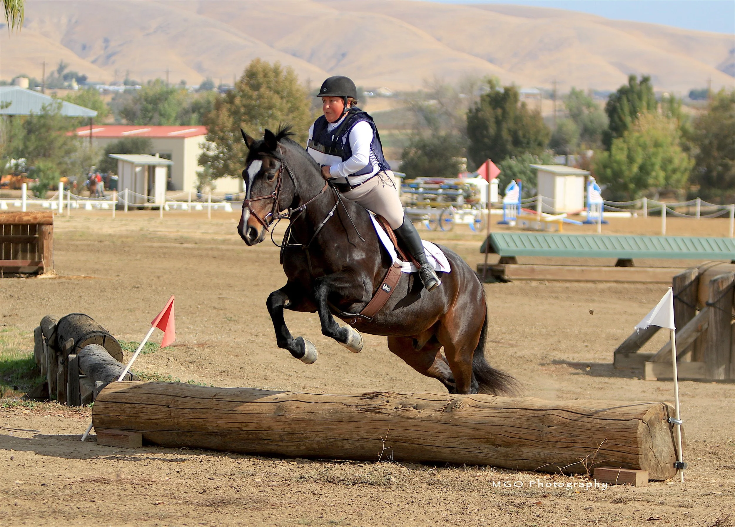 A person riding a horse competes in an equestrian event, jumping over a log obstacle on a dirt course with mountains and tents visible in the background.
