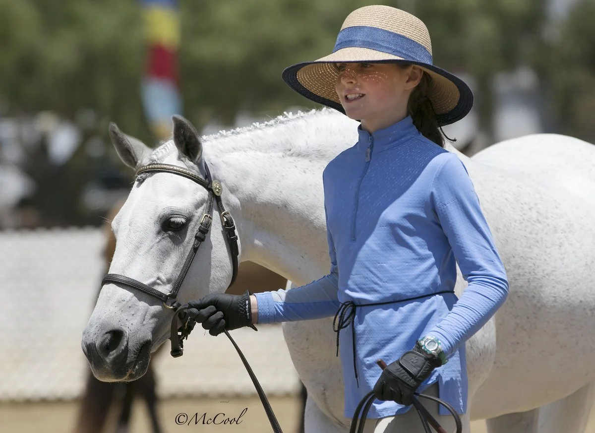 A girl dressed in blue, wearing a sun hat and riding gloves, stands next to a white horse, holding its bridle, in an outdoor setting.