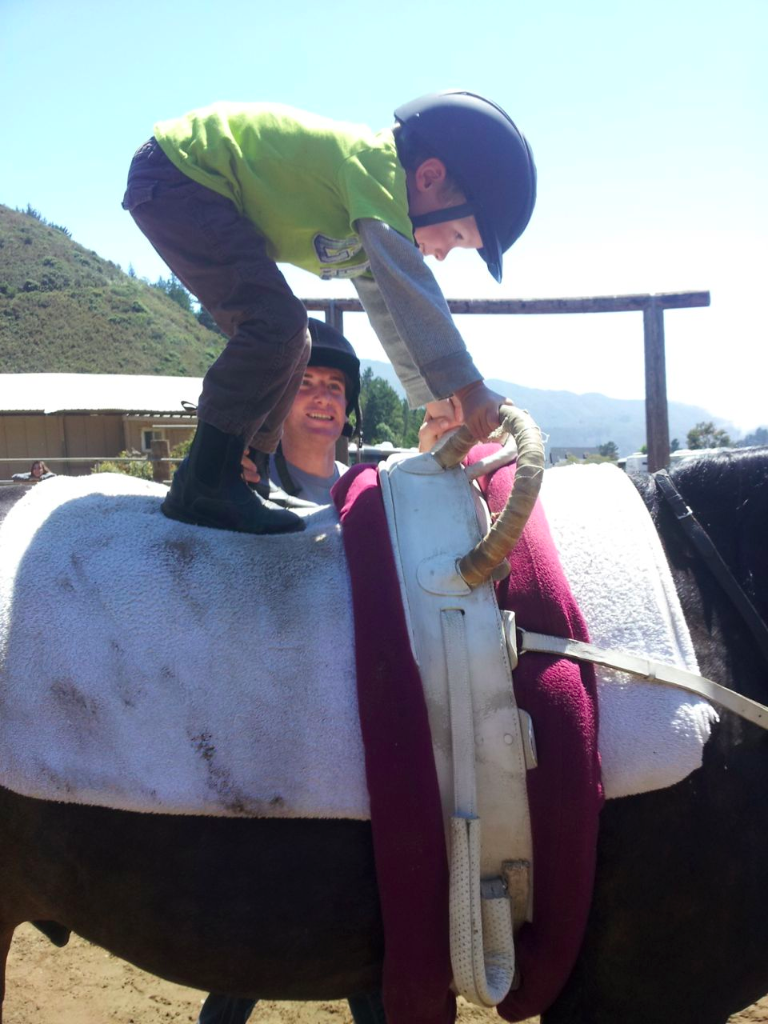A young boy in a green shirt and black helmet is riding on a saddle on a horse, with a woman behind him guiding the horse. The man is wearing a helmet and a blue shirt.
