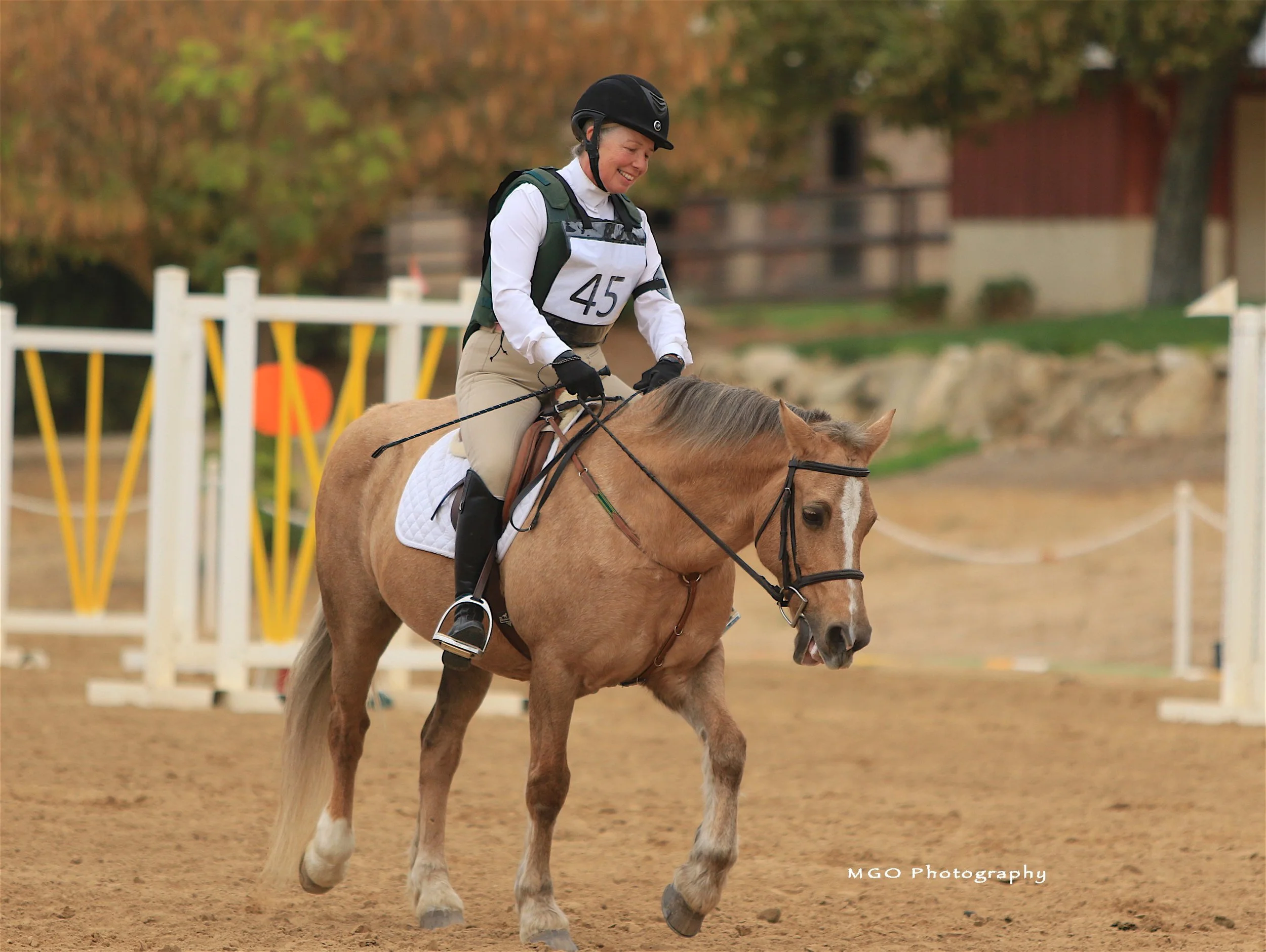 A smiling woman wearing a black helmet, white long-sleeve shirt, and beige riding pants riding a light brown horse with a white blaze on its face during a horseback riding competition. She is holding the reins as the horse walks on a dirt arena with 