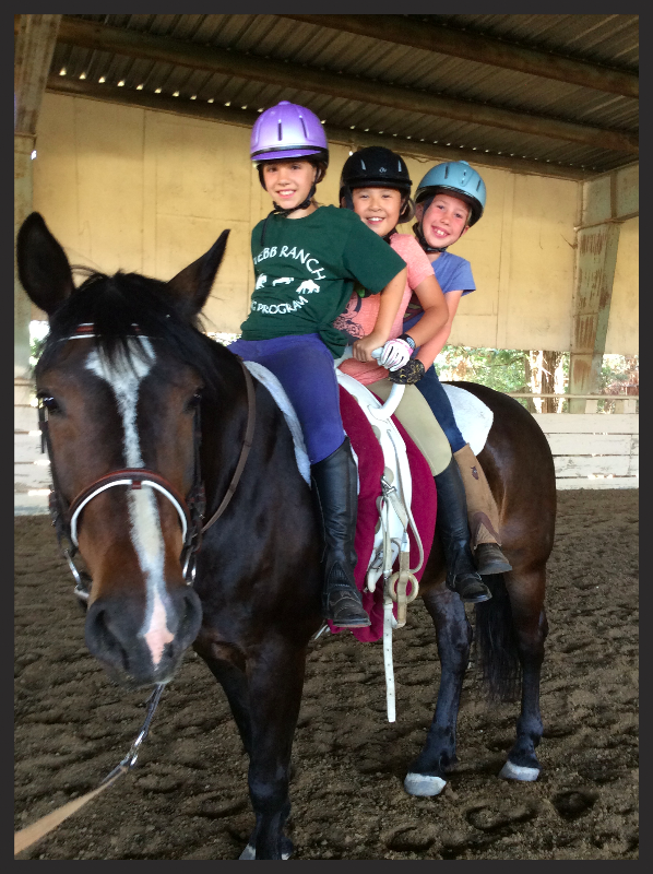 Three children wearing helmets smiling while riding a horse inside an indoor riding arena.