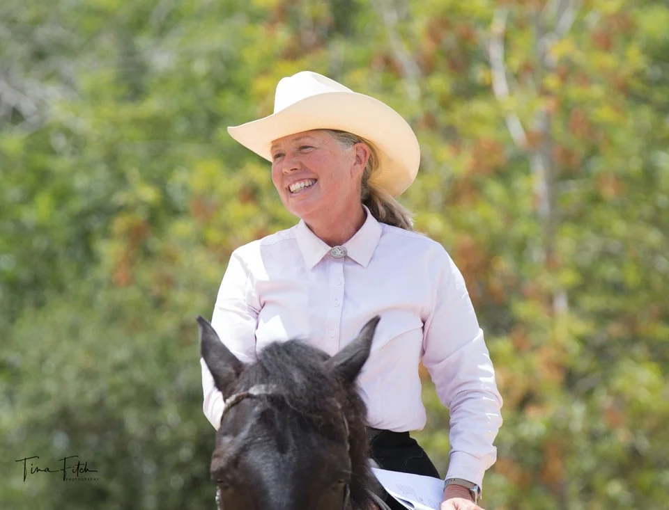 A woman with a white cowboy hat and white shirt riding a black horse outdoors, smiling amidst fall foliage.