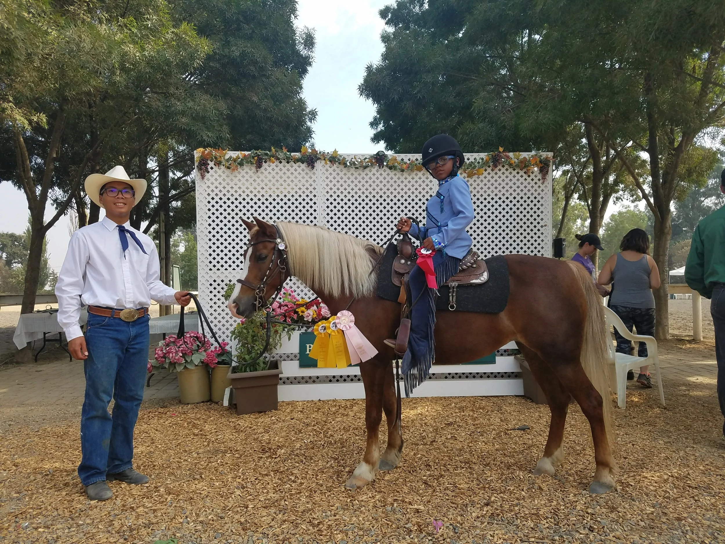 Young boy in a light blue shirt, dark jeans, and a black helmet riding a brown pony with a blonde mane. A man in western attire, including a white cowboy hat, white shirt, and blue jeans, stands beside the pony holding its lead rope. The background f