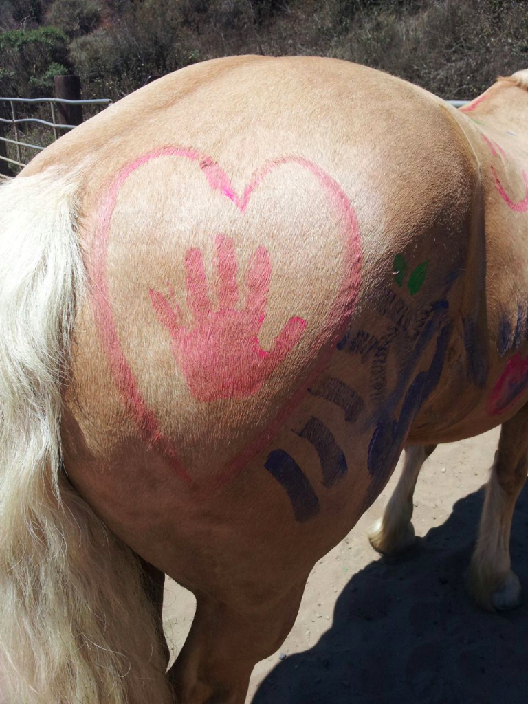 A light-colored horse with red, green, and black painted designs on its side, including a heart with a handprint inside it.