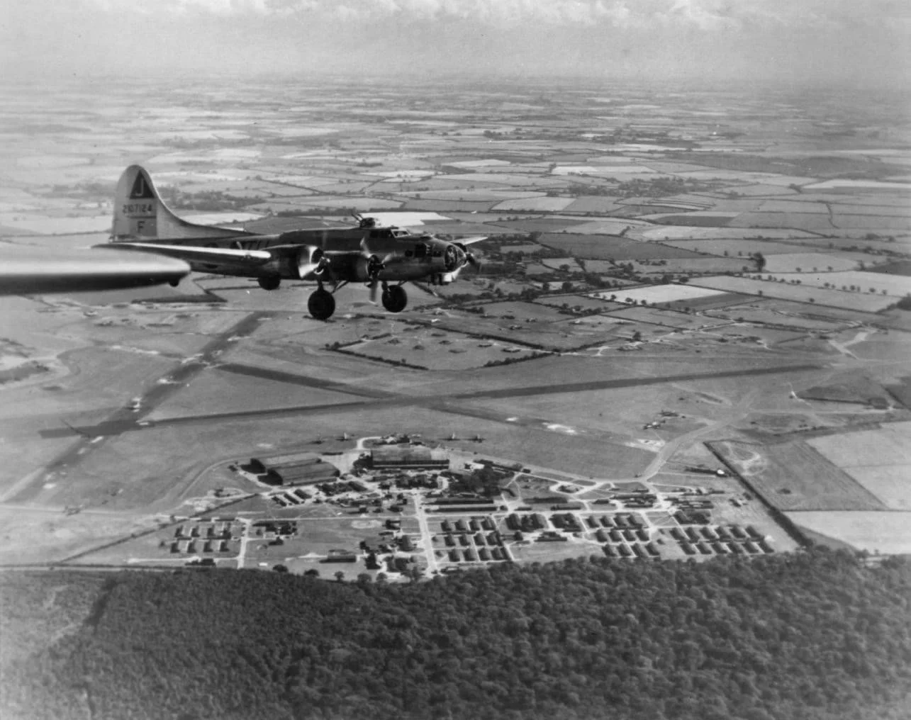 Black and white aerial photograph showing a military aircraft flying over a rural landscape with fields, roads, and a small village.